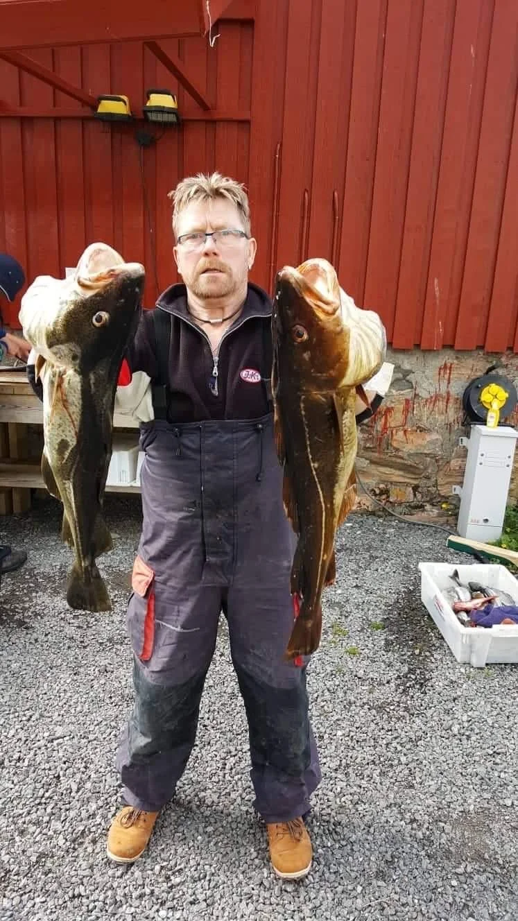 Ein Mann steht vor einer roten Holzwand und hält zwei große, gefangene Fische, einen in jeder Hand.