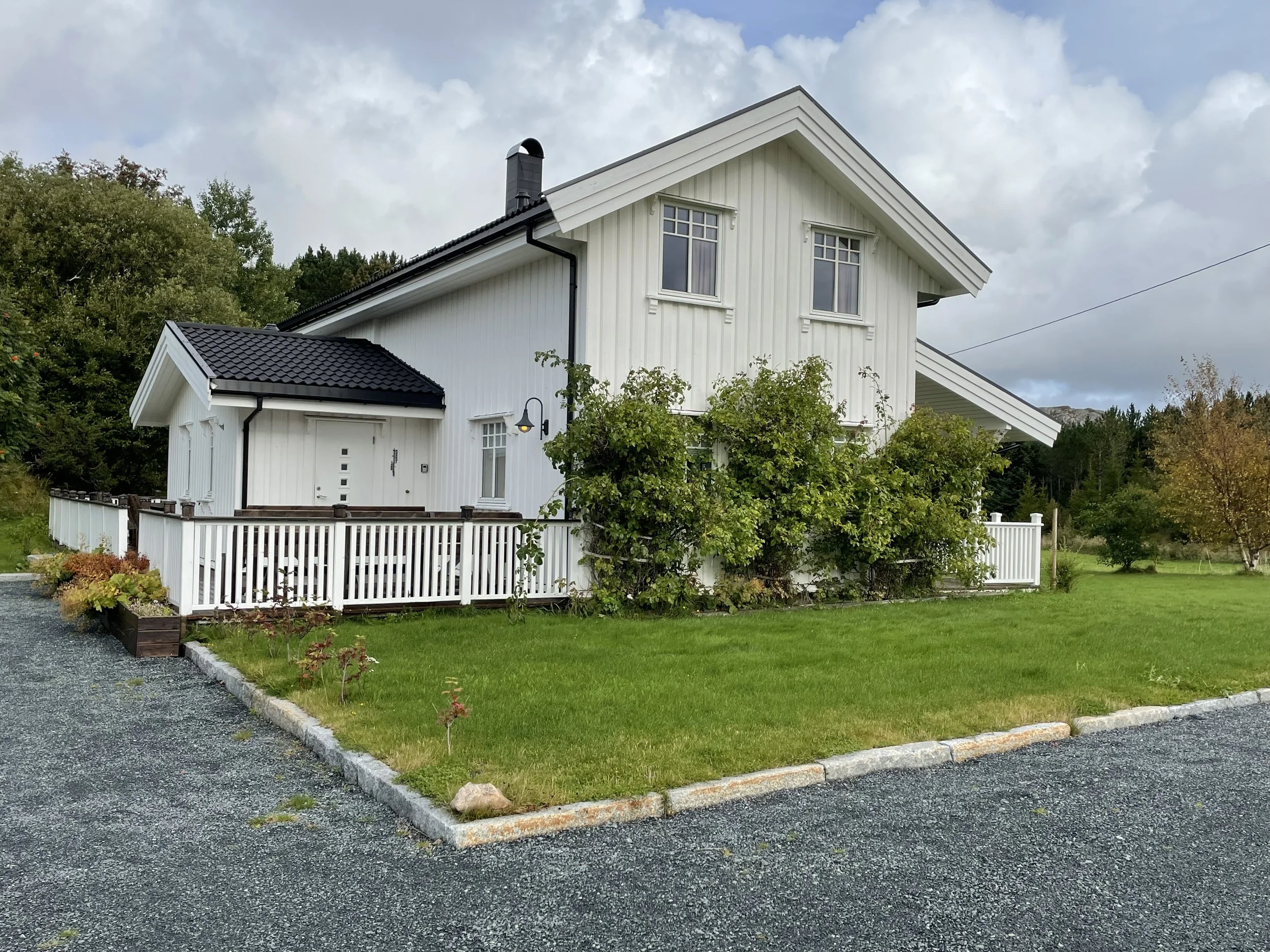 Ein weißes, zweistöckiges Haus mit einem schwarzen Dach und mehreren Fenstern, umgeben von grüner Vegetation und einem bewölkten Himmel.