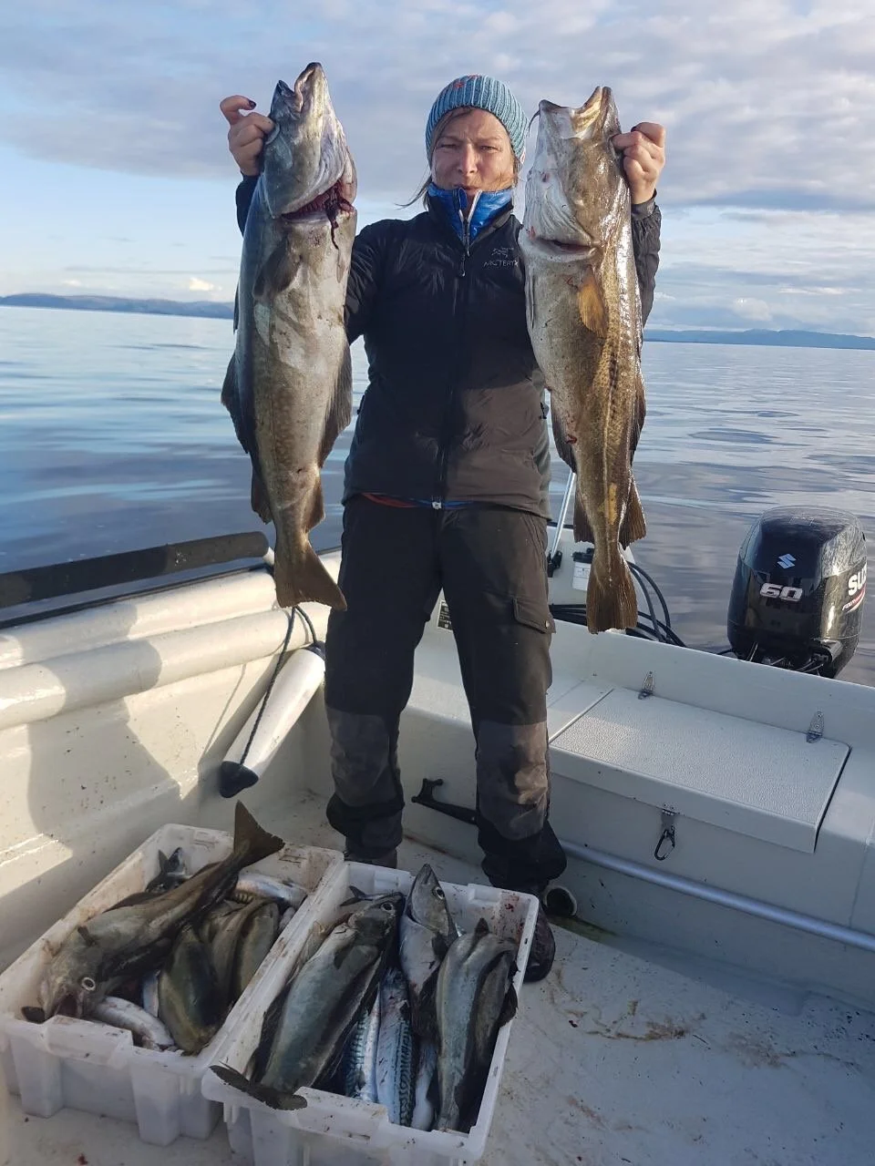 Eine Frau auf einem Boot, die zwei große Fische hält, mit einem offenen Kasten voller gefangener Fische, im Hintergrund ein ruhiges Gewässer und ein bewölkter Himmel.