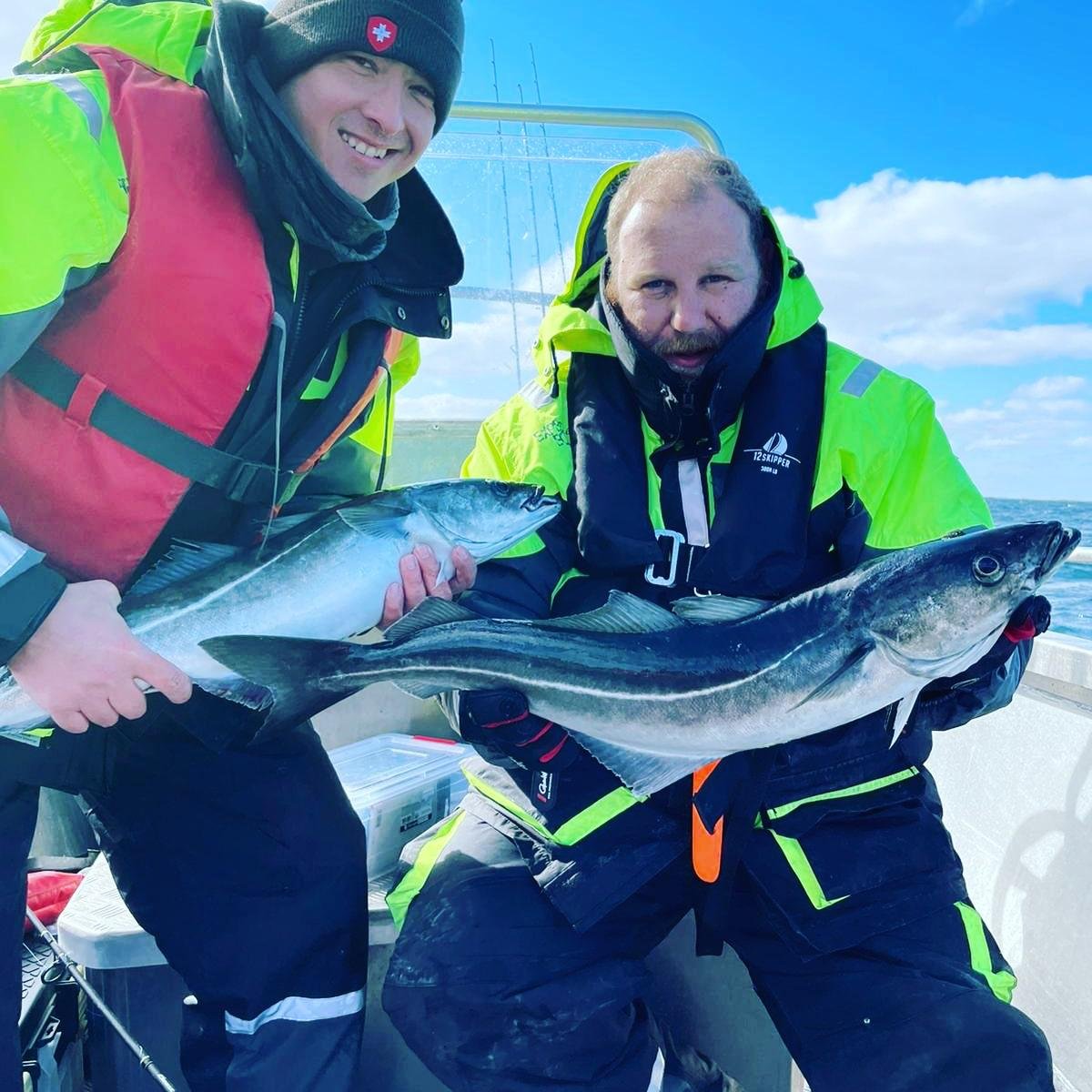 Zwei Fischer auf einem Boot, die jeweils große Fische gehalten, mit Blick auf das Meer und Himmel im Hintergrund.