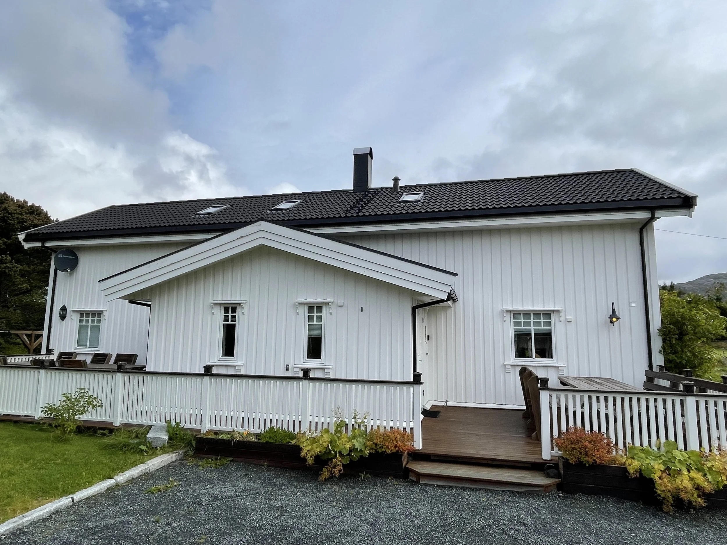 Ein weißes Haus mit schwarzen Ziegeldach, Holztreppe, Garten, Holzdekor und mehreren Fenstern, umgeben von grünem Garten und Bäumen, unter bewölktem Himmel.