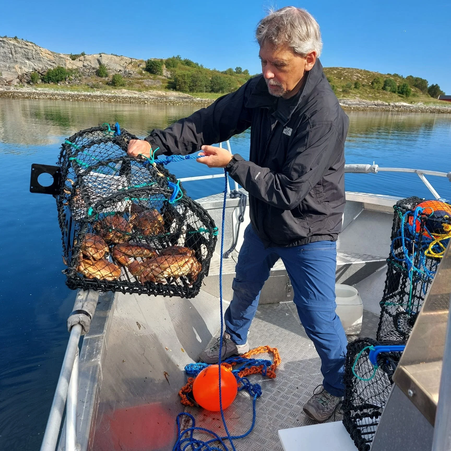 Mann auf einem Boot beim Krabbenfang mit Netz und Krabbenkörben auf ruhigem Wasser, im Hintergrund Berge und Himmel.