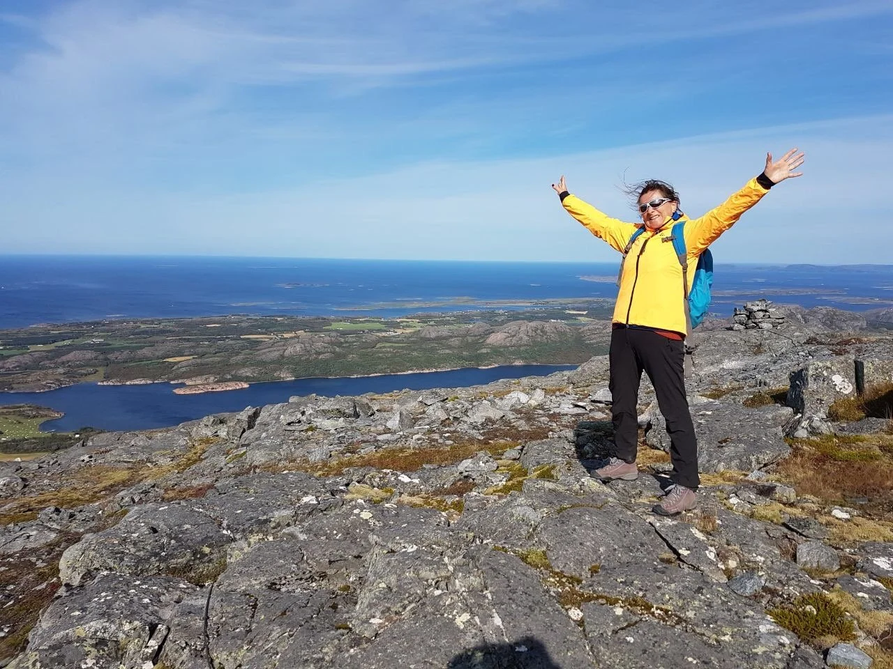 Eine Frau in gelber Jacke und schwarzen Hosen steht auf einem Felsen auf einem Berggipfel mit ausgestreckten Armen, im Hintergrund diagonal verlaufende Seen, grüne Felder, Meeresküste und blauer Himmel mit wenigen Wolken.