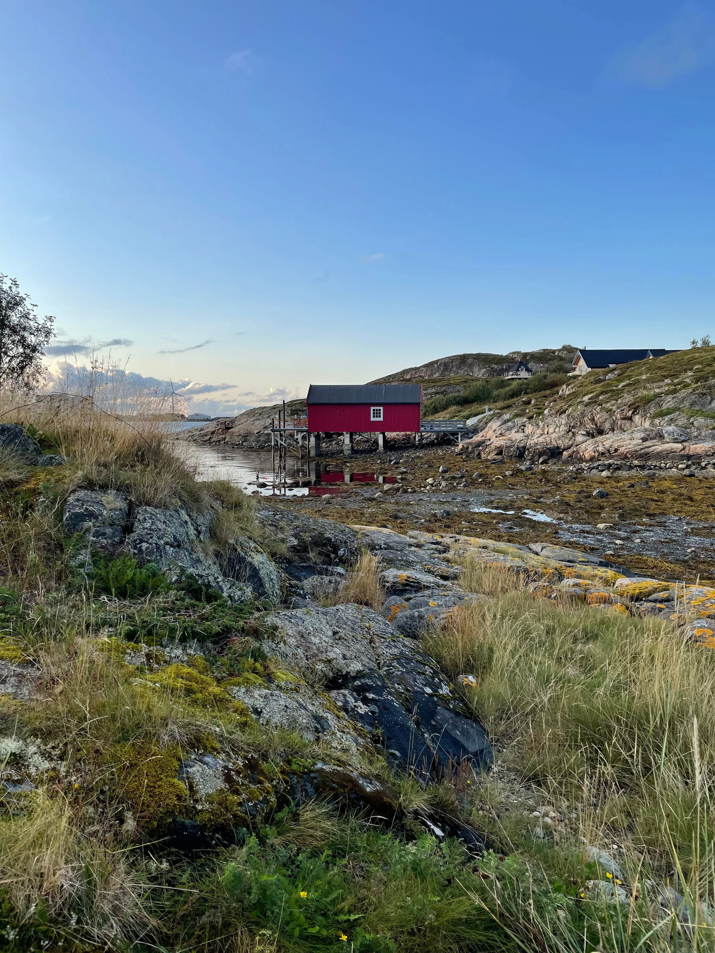 Ein rotes Haus auf Stelzen am Wasser in einer felsigen, grasbedeckten Küstenlandschaft bei Sonnenuntergang.