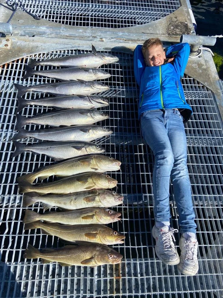 Ein Junge liegt lachend auf einem Metallgitterboden neben einer Reihe gefangener Fische an einem Bootssteg.