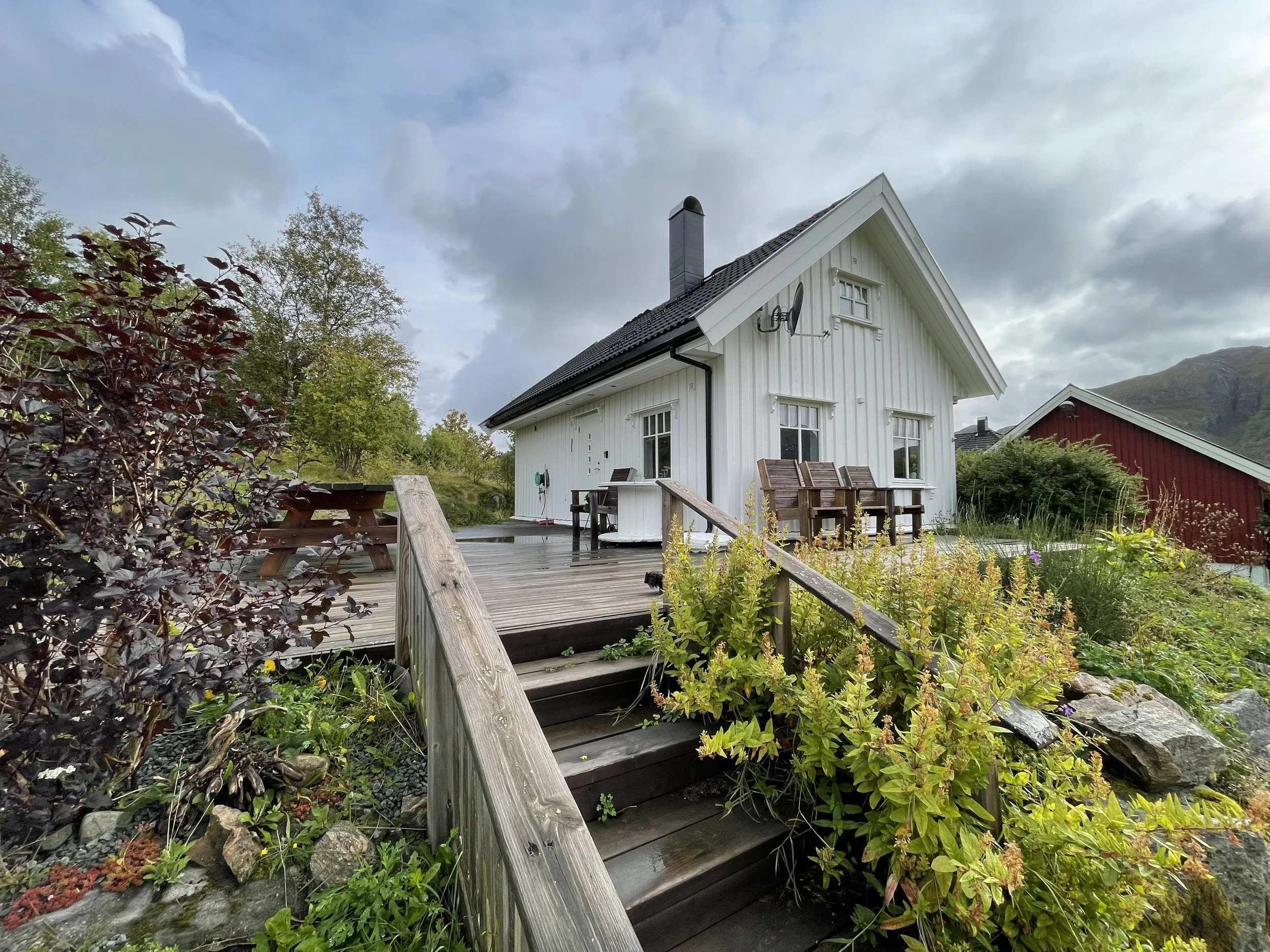 Ein weißes Holzhaus mit einem aufgeschobenen Holzdeck und Gartenmöbeln, umgeben von Pflanzen und Bäumen, unter einem bewölkten Himmel in einer bergigen Landschaft.