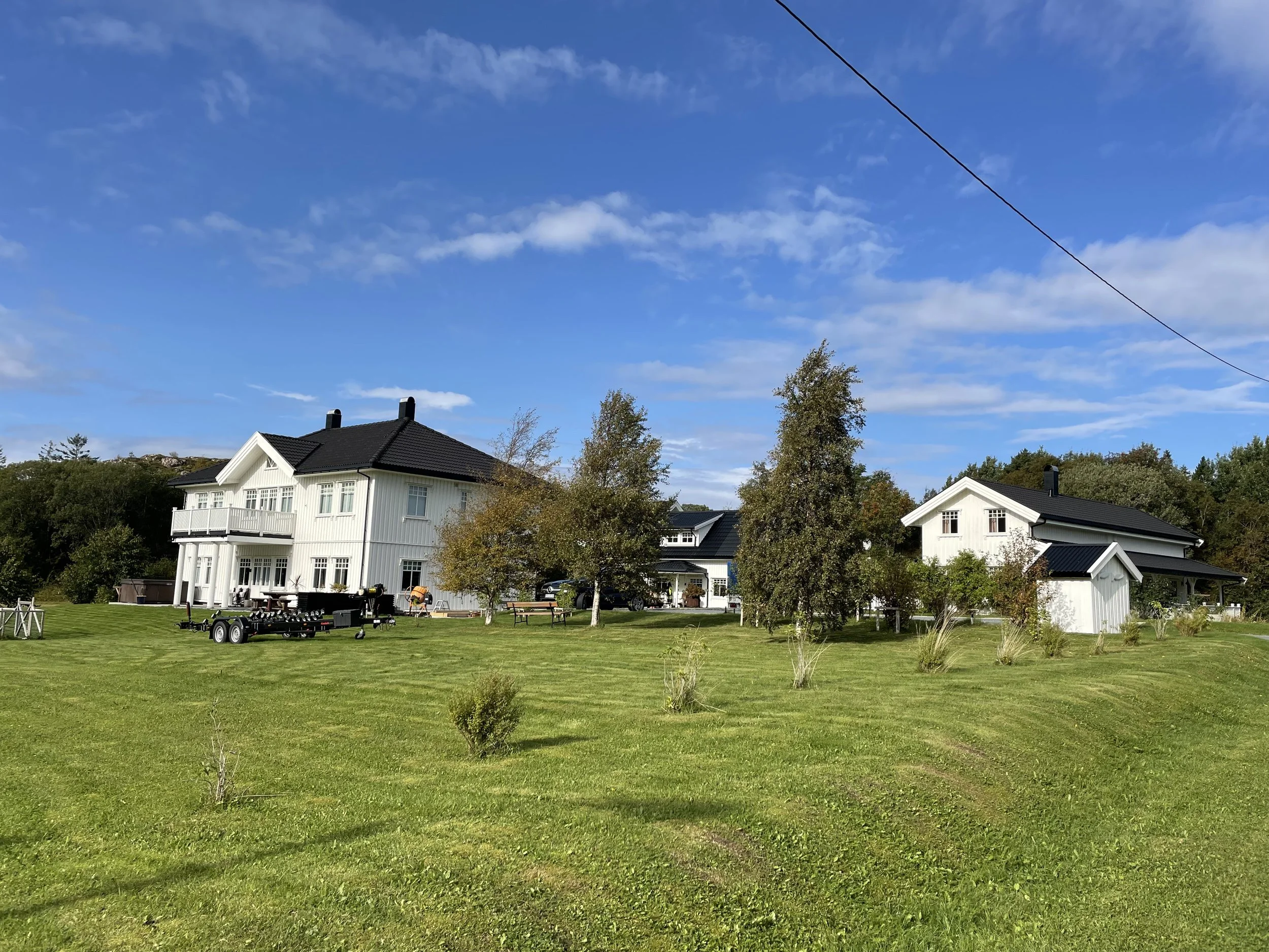 Ein weißes Haus mit schwarzem Dach auf einer grünen Wiese, umgeben von Bäumen, bei sonnigem Himmel.