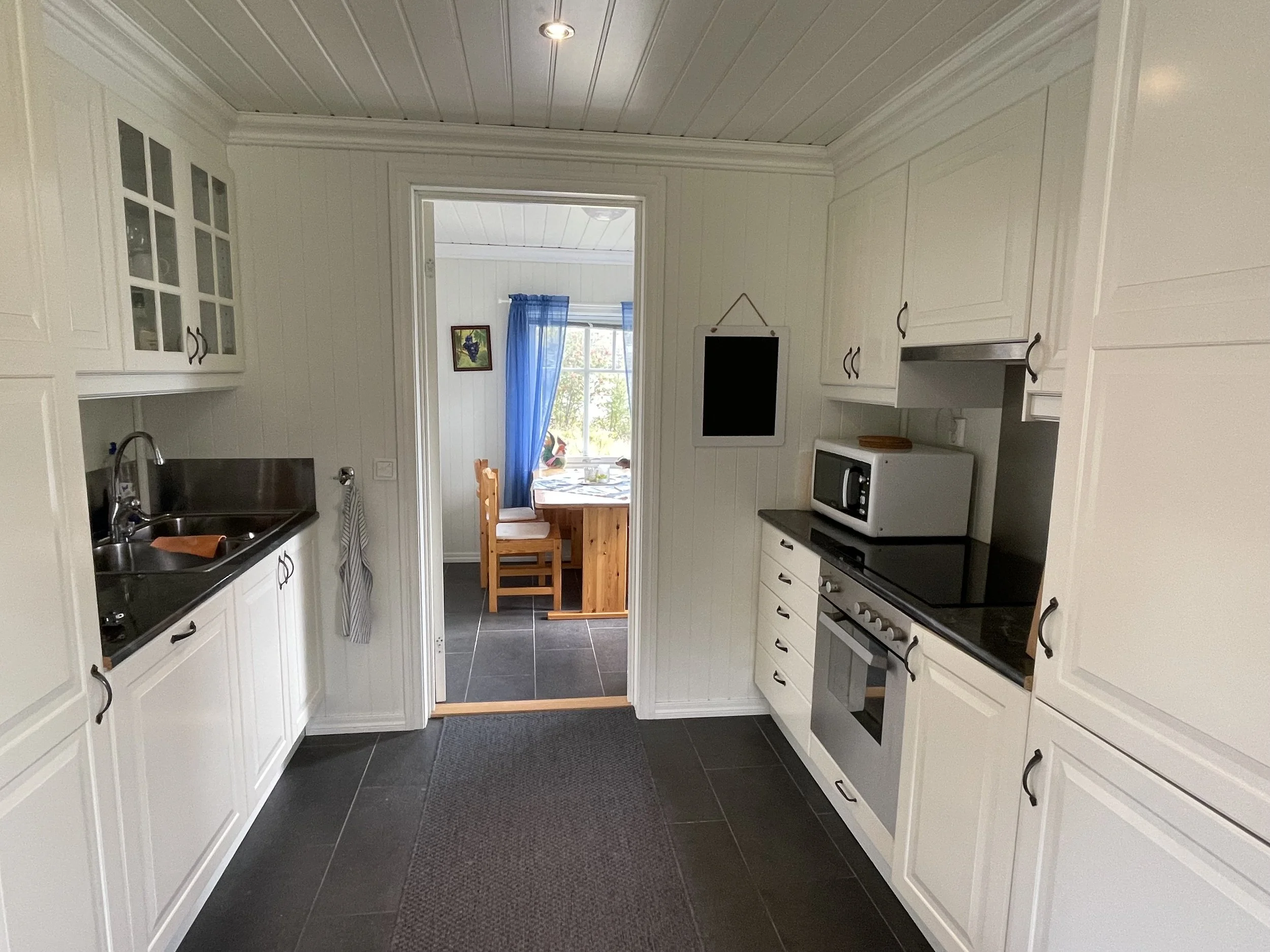 Innenansicht einer modernen Küche mit weißen Schränken, schwarzen Armaturen, Herd, Mikrowelle und einem Esstisch im Hintergrund mit blauen Vorhängen am Fenster.