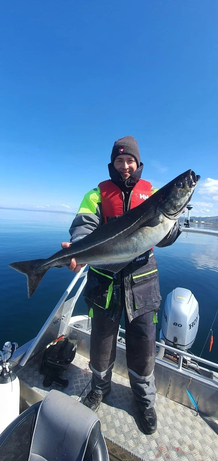 Mann auf dem Boot hält einen großen Fisch vor blauem Himmel und ruhigem Wasser.