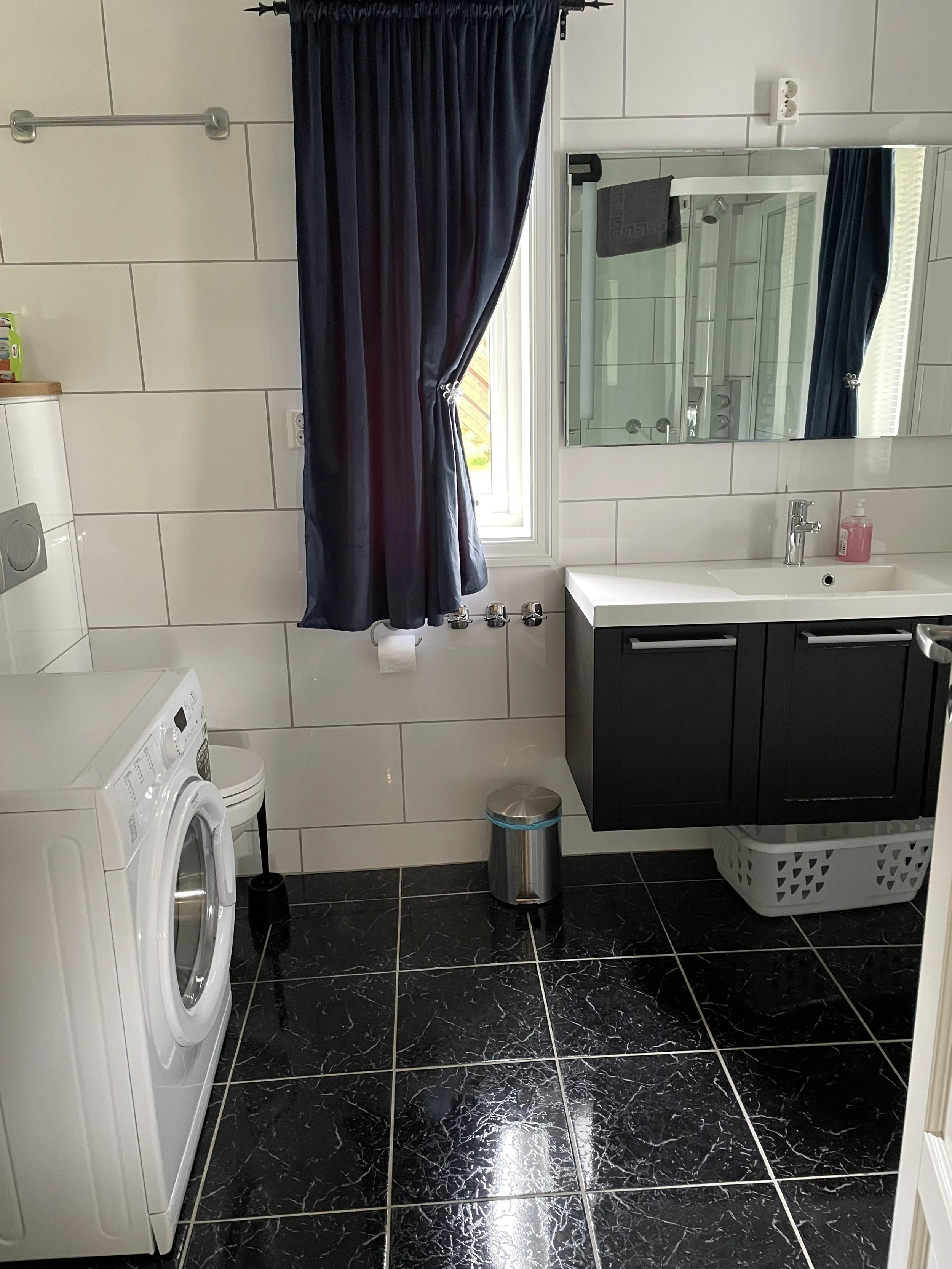 Ein Badezimmer mit weißen Wandfliesen, schwarzem Boden mit weißen Marmorstreifen, einer Waschmaschine, einem Waschbecken mit Spiegel, einem Handtuch, einem Mülleimer, einem Badezimmerspiegel und einem Fenster mit dunklen Vorhängen.