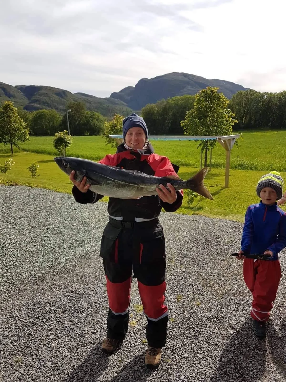 Eine Person hält einen großen Fisch vor einer grünen, bewaldeten Landschaft mit Bergen im Hintergrund, während ein Kind neben ihr steht.