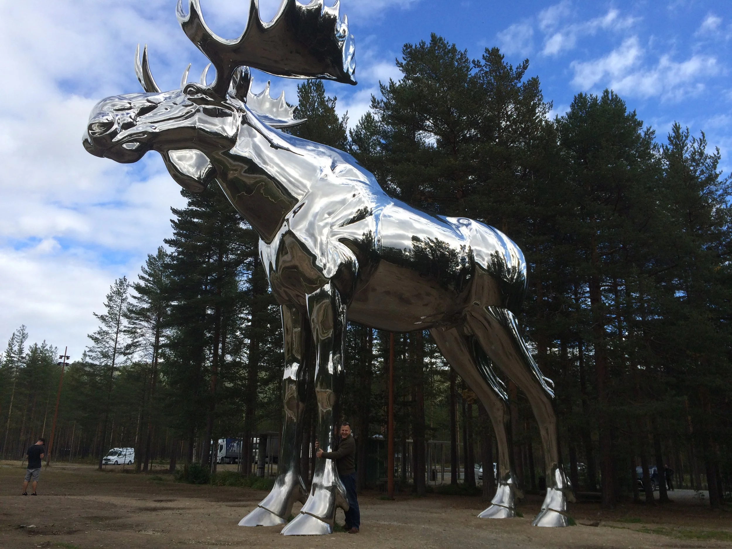 Große, glänzende metallenes Reh-Statue im Freien vor einem Himmel mit Wolken und Bäumen. Eine Person steht neben der Statue.