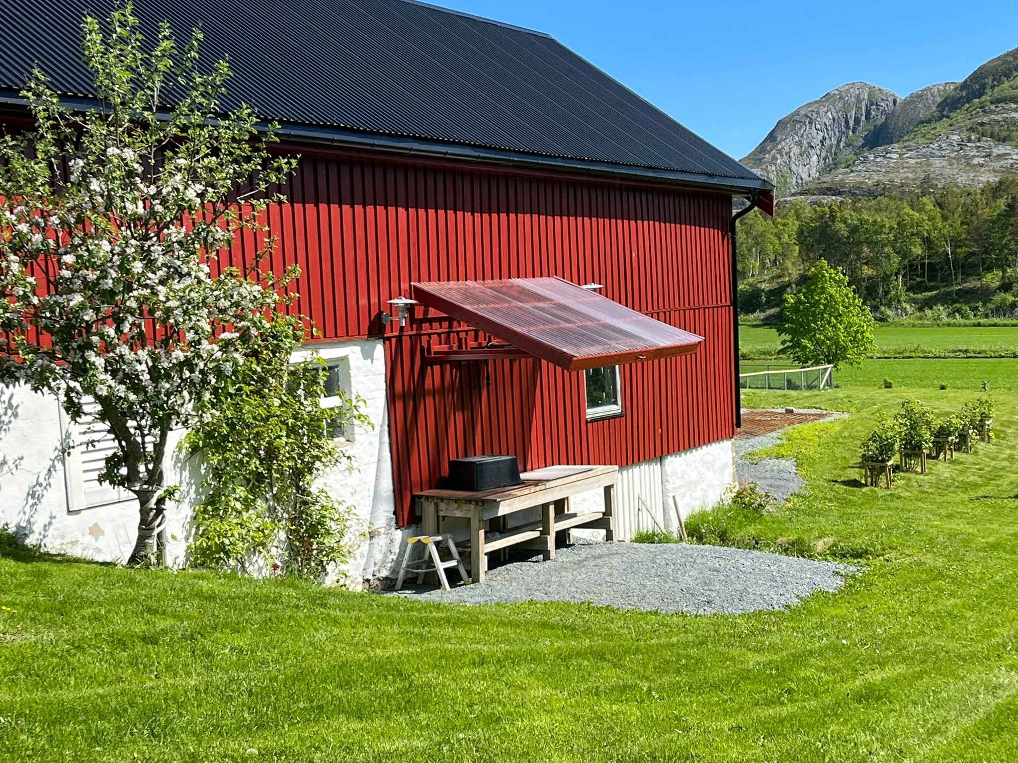 Ein rotes Bauernhaus mit schwarzem Dach und weißem unteren Bereich, umgeben von grünen Wiesen und Bäumen, im Hintergrund Berge und blauer Himmel.