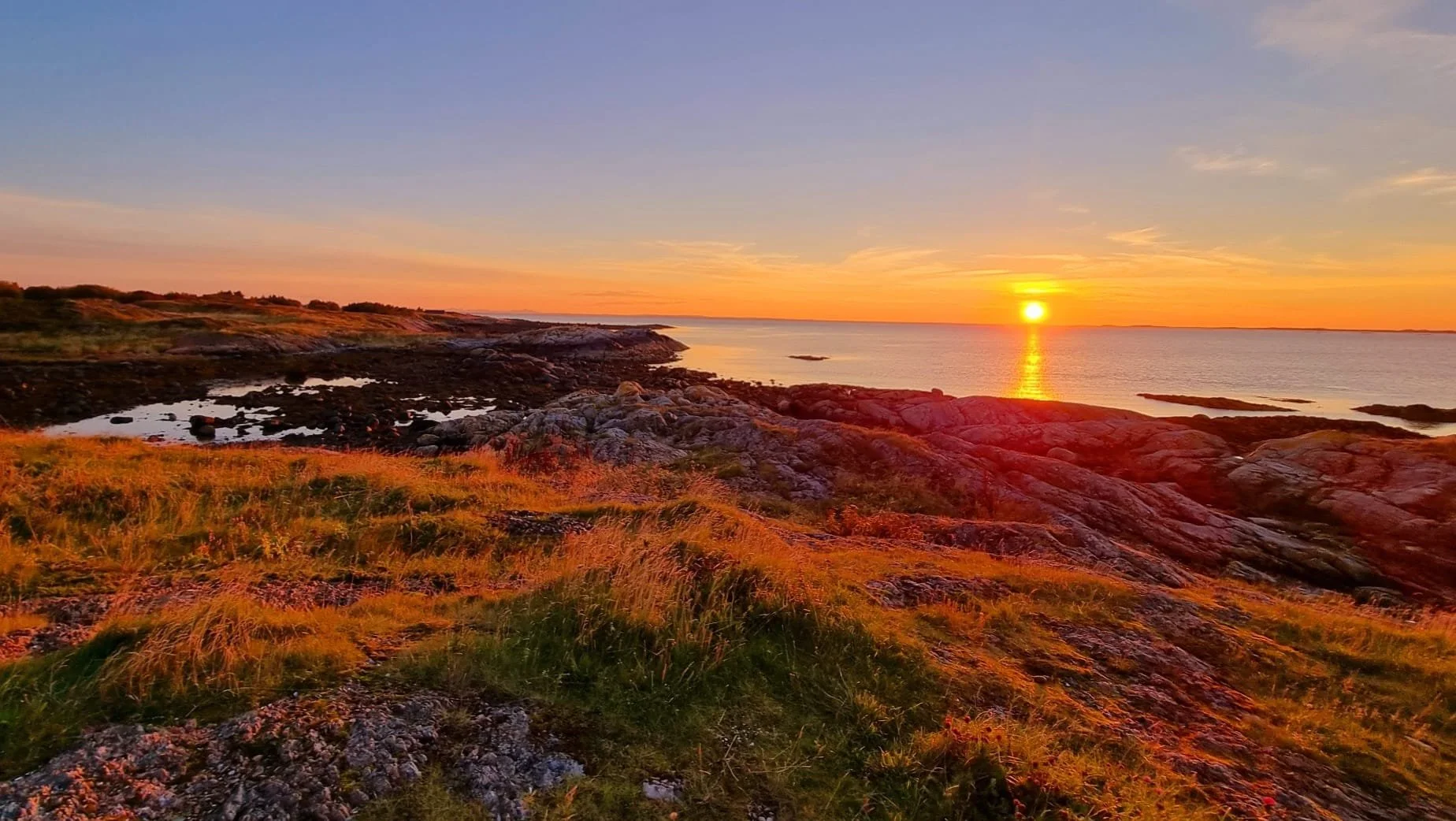 Ein Sonnenuntergang über einer felsigen Küstenlandschaft mit grünem Gras und kleinen Wasserlachen.