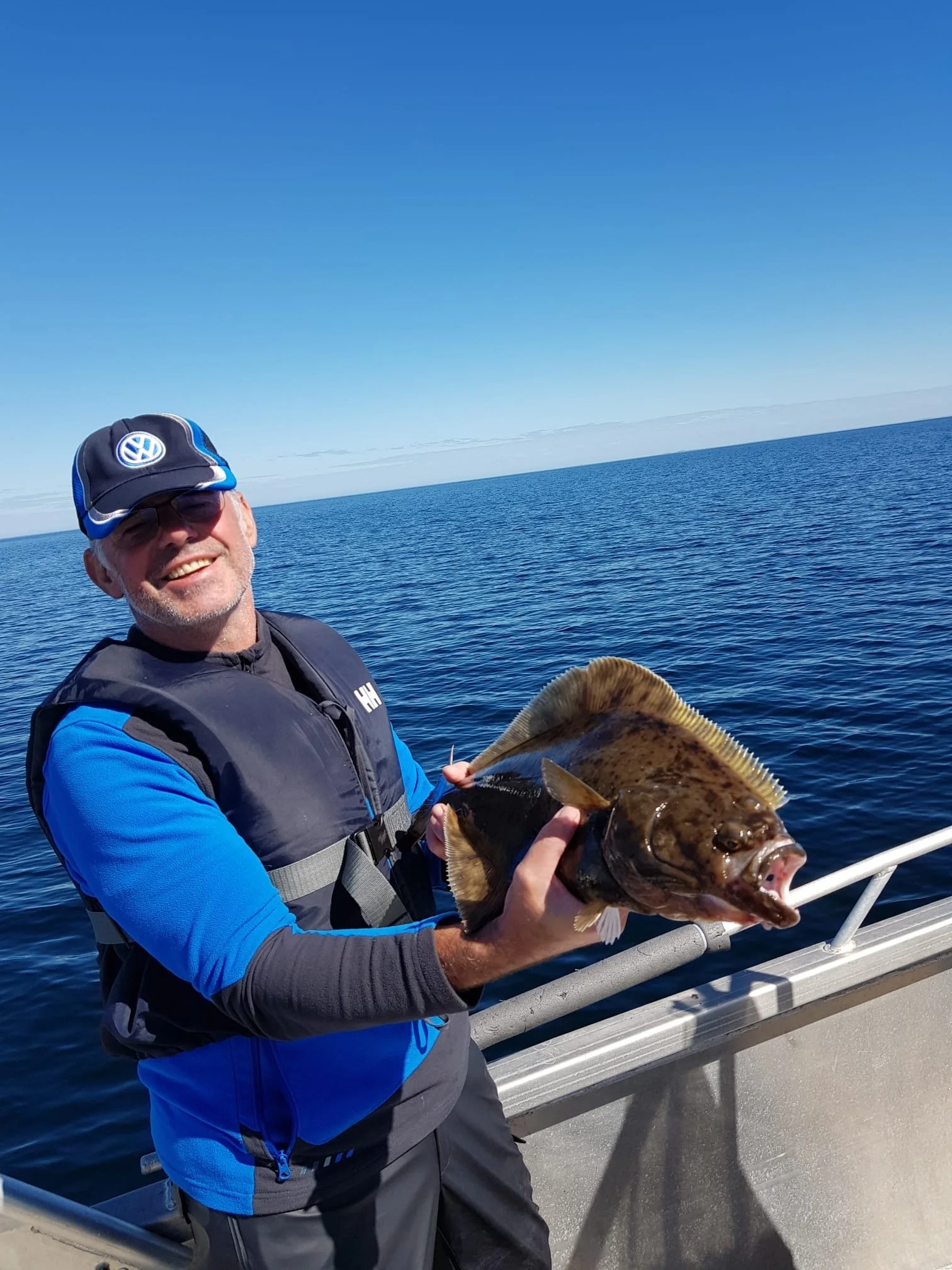 Mann auf einem Boot hält einen großen Fisch in der Hand, blauer Himmel und ruhiges Meer im Hintergrund.