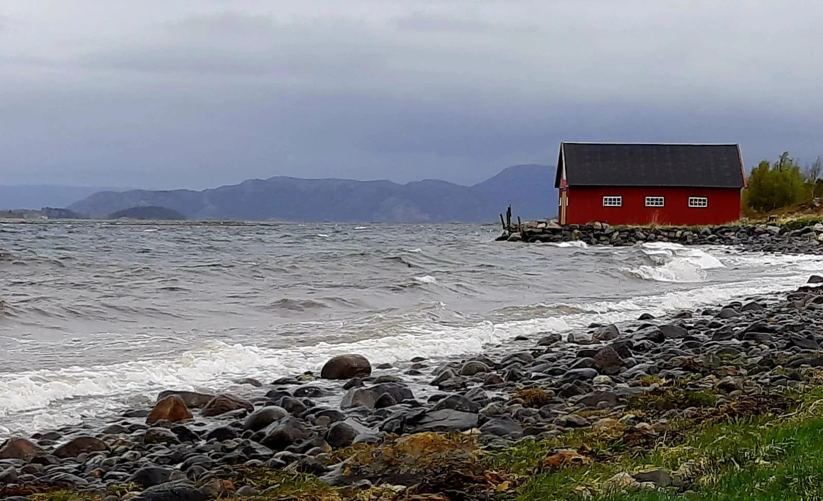 Küstendorf mit einem roten Haus am Ufer, Wellen, Steine und Berge im Hintergrund an einem bewölkten Tag.