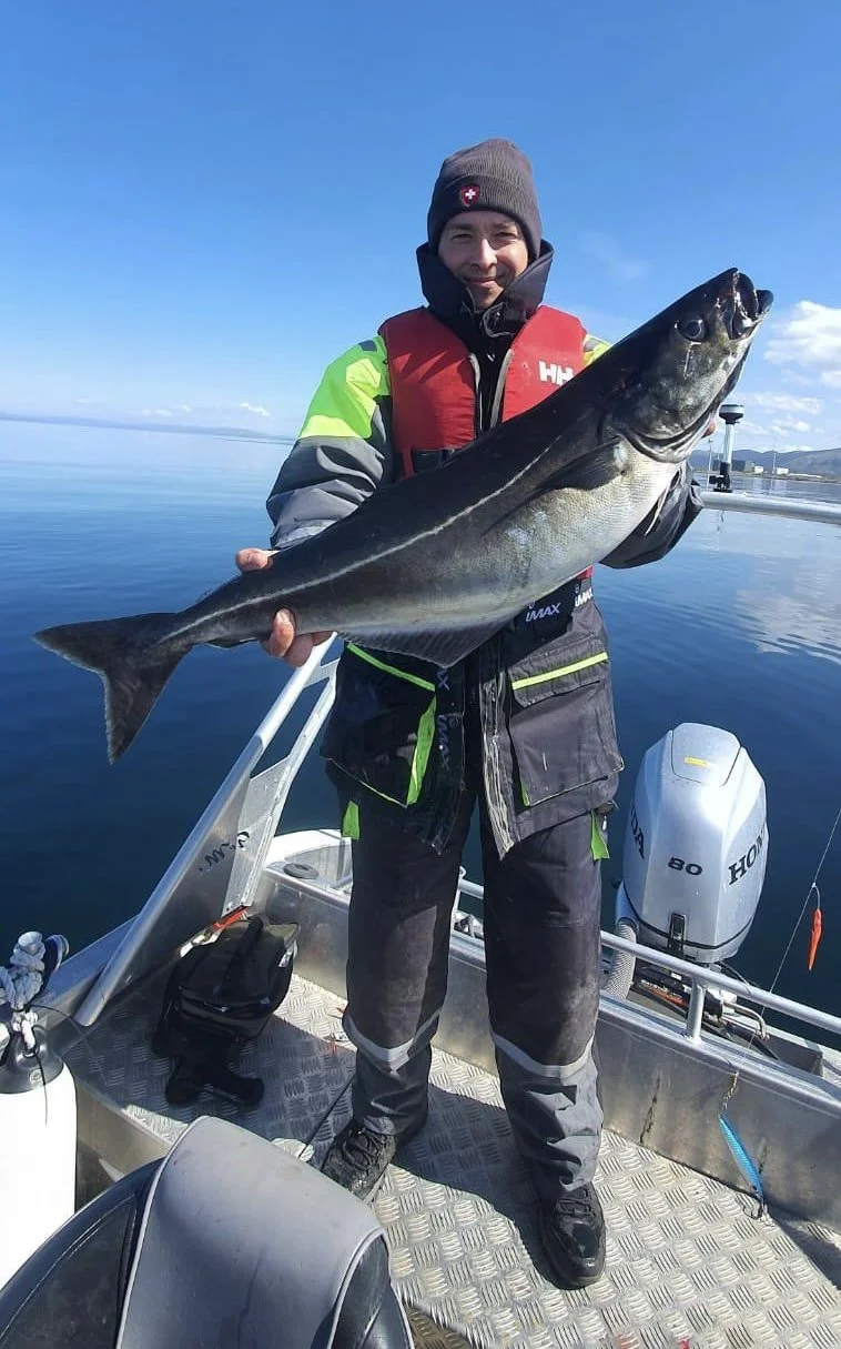 Mann auf einem Boot hält einen großen Fisch vor Wasser mit Himmel im Hintergrund, trägt Winterkleidung und eine rote Schwimmweste.
