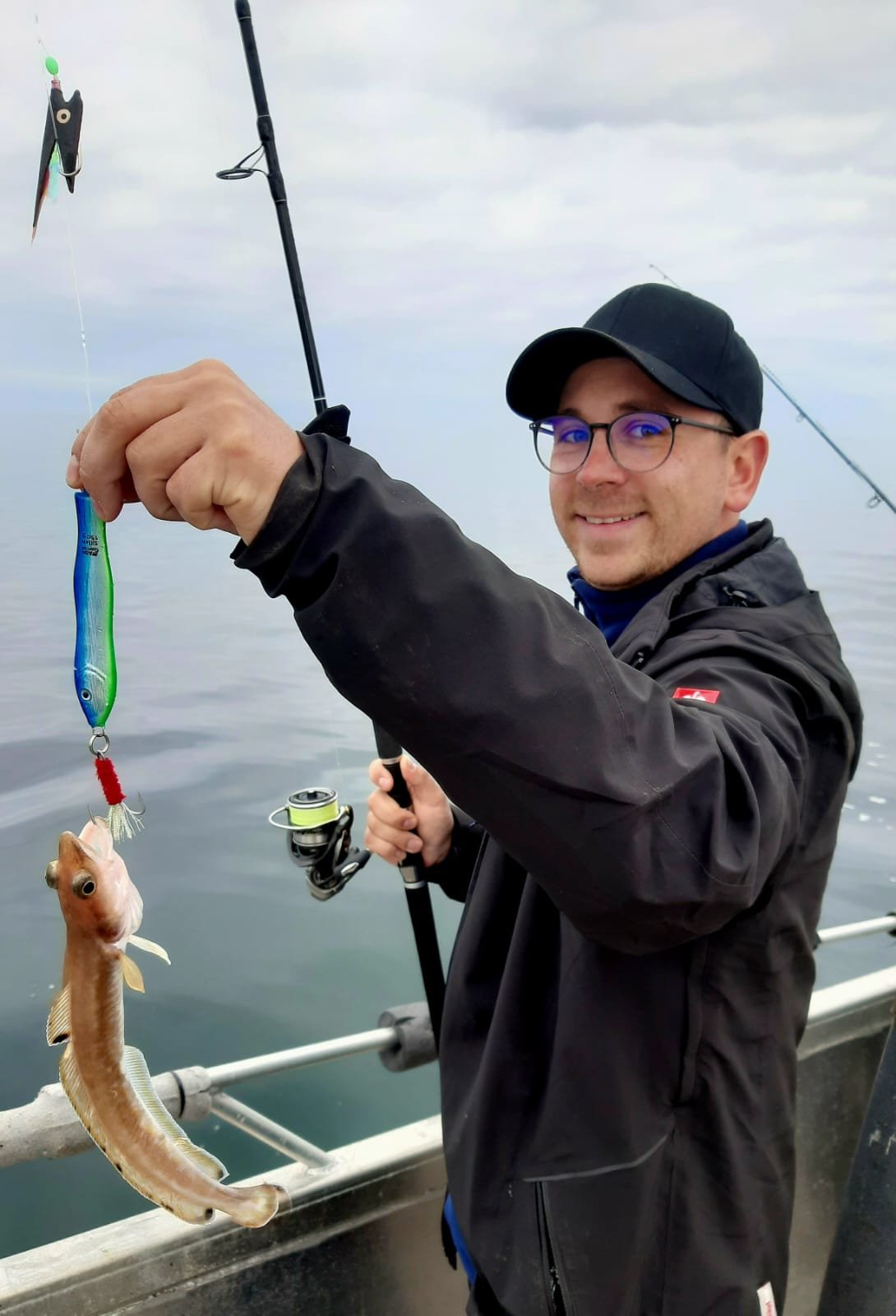Mann mit Brille und schwarzem Hut hält einen Fisch, der an einem Angelhaken hängt, während er auf einem Boot im Wasser steht.