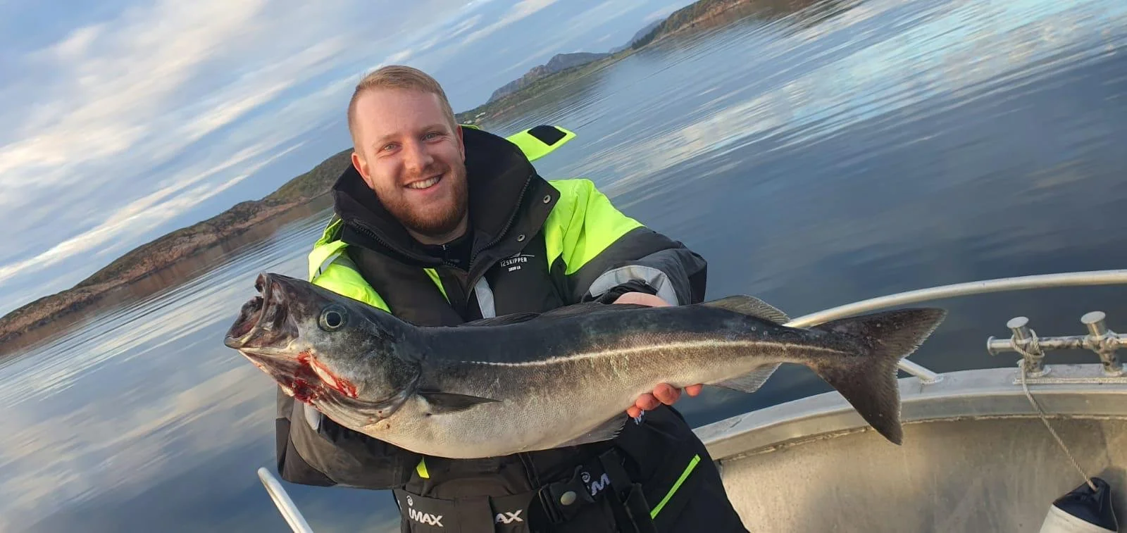 Ein Mann auf einem Boot hält einen großen Fisch in den Händen, während er auf dem Wasser sitzt, mit Bergen im Hintergrund.