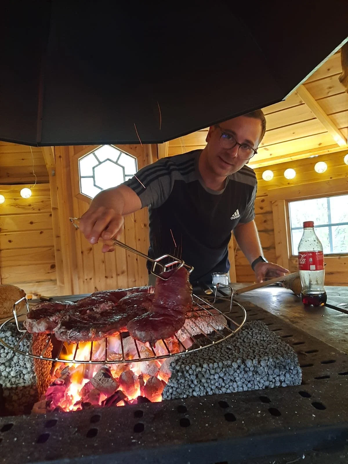 Ein Mann grillt Fleisch über einem Holzkohlegrill in einem Holzraum, neben ihm steht eine Flasche Cola.
