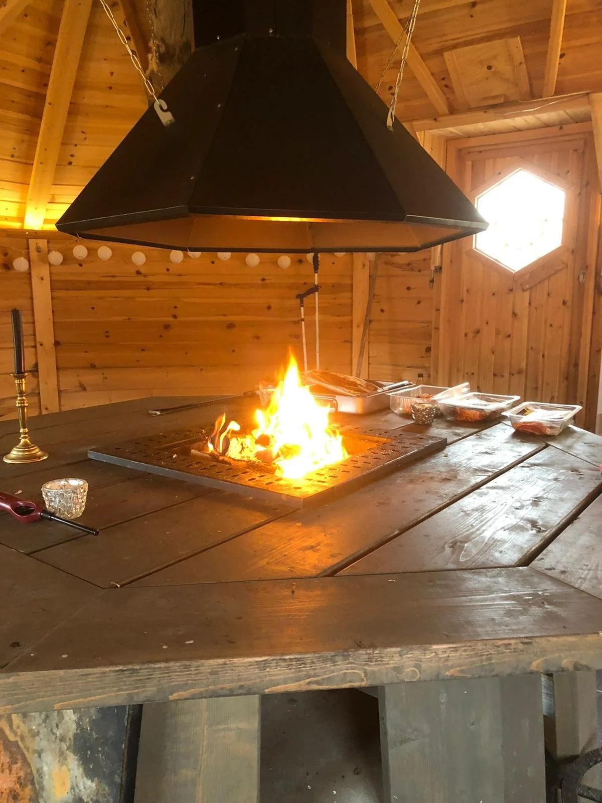 Ein Tisch mit offenem Feuer in einer hölzernen Hütte, umgeben von Tellern und Gewürzen, mit einem großen schwarzen Hängeschirm darüber.