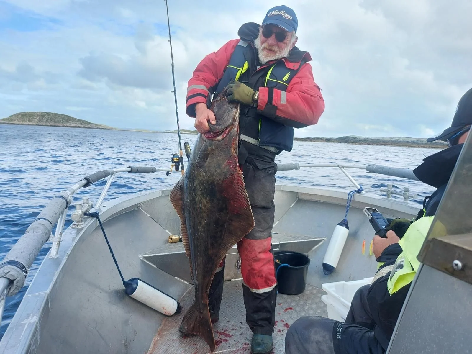 Ein älterer Mann mit Sonnenbrille, Polizeiklamotten und roter Jacke hält einen großen Fisch auf einem Boot im Meer. Im Hintergrund sind kleine Inseln und eine bewölkte Himmel zu sehen.