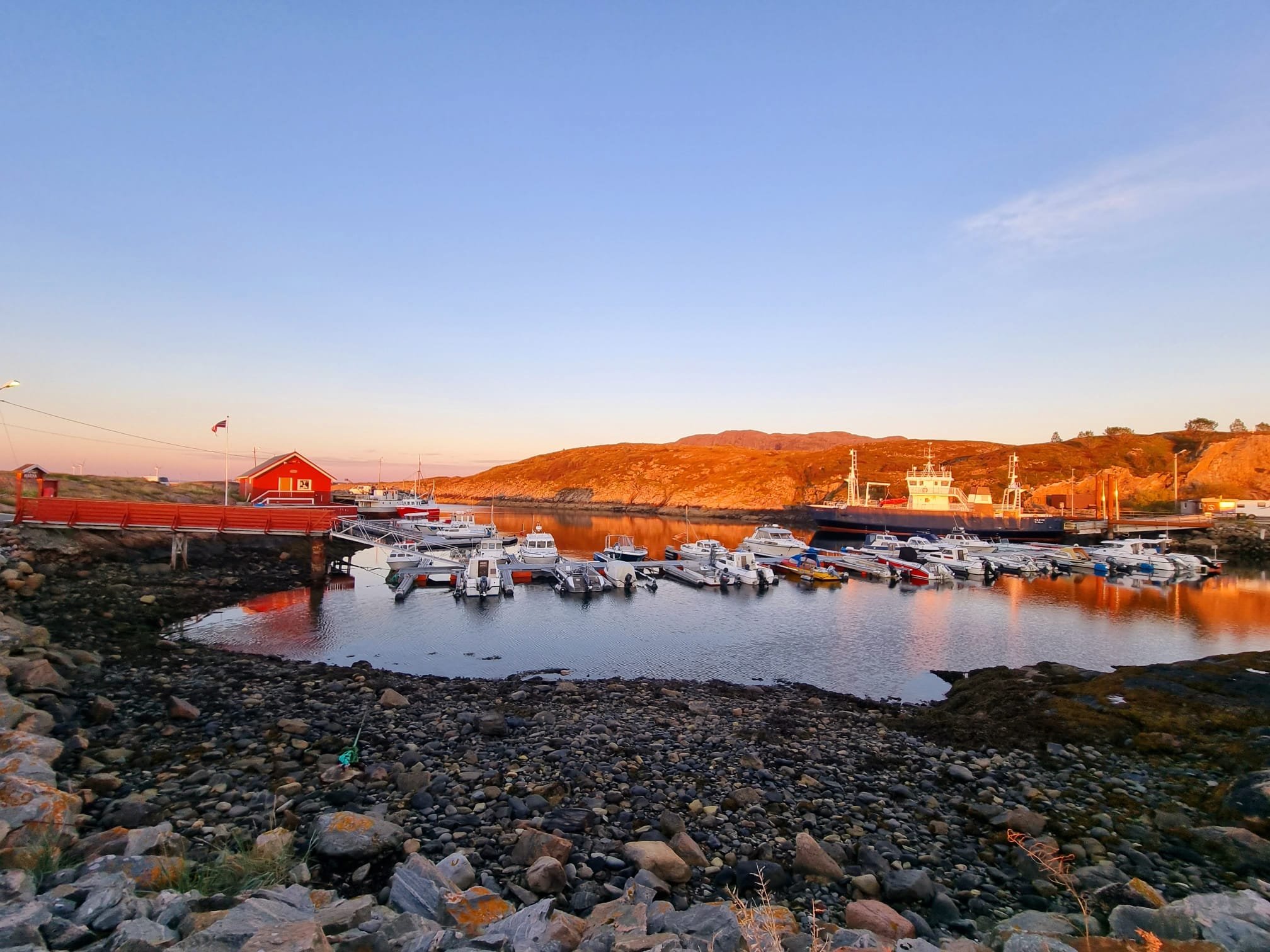Ein Hafen mit mehreren Booten, einem roten Gebäude und einer Felsenlandschaft bei Sonnenuntergang.
