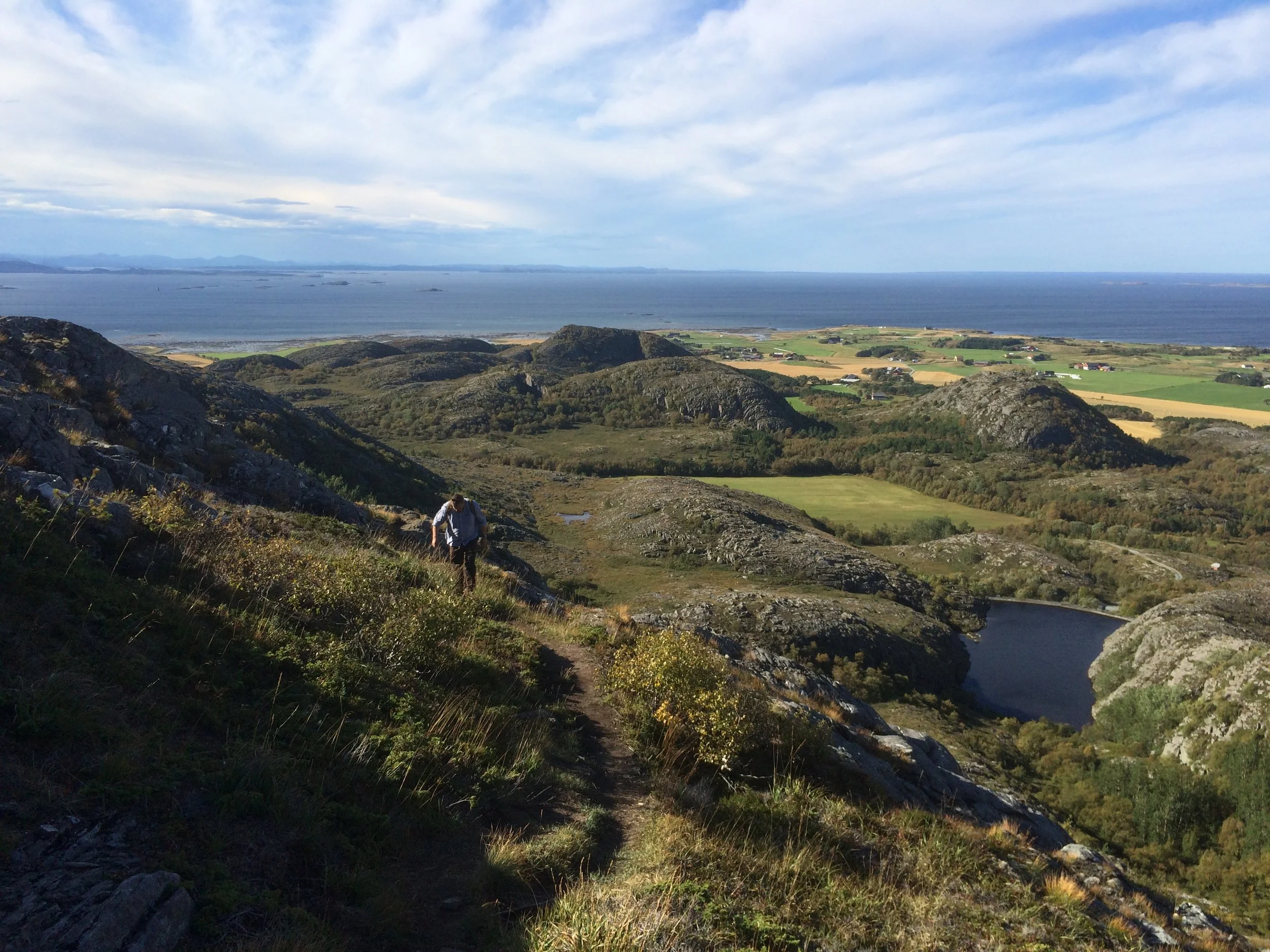 Ein Wanderer auf einem Bergpfad mit Blick auf eine grüne Landschaft, Seen, Berge, das Meer und Wolken am Himmel.