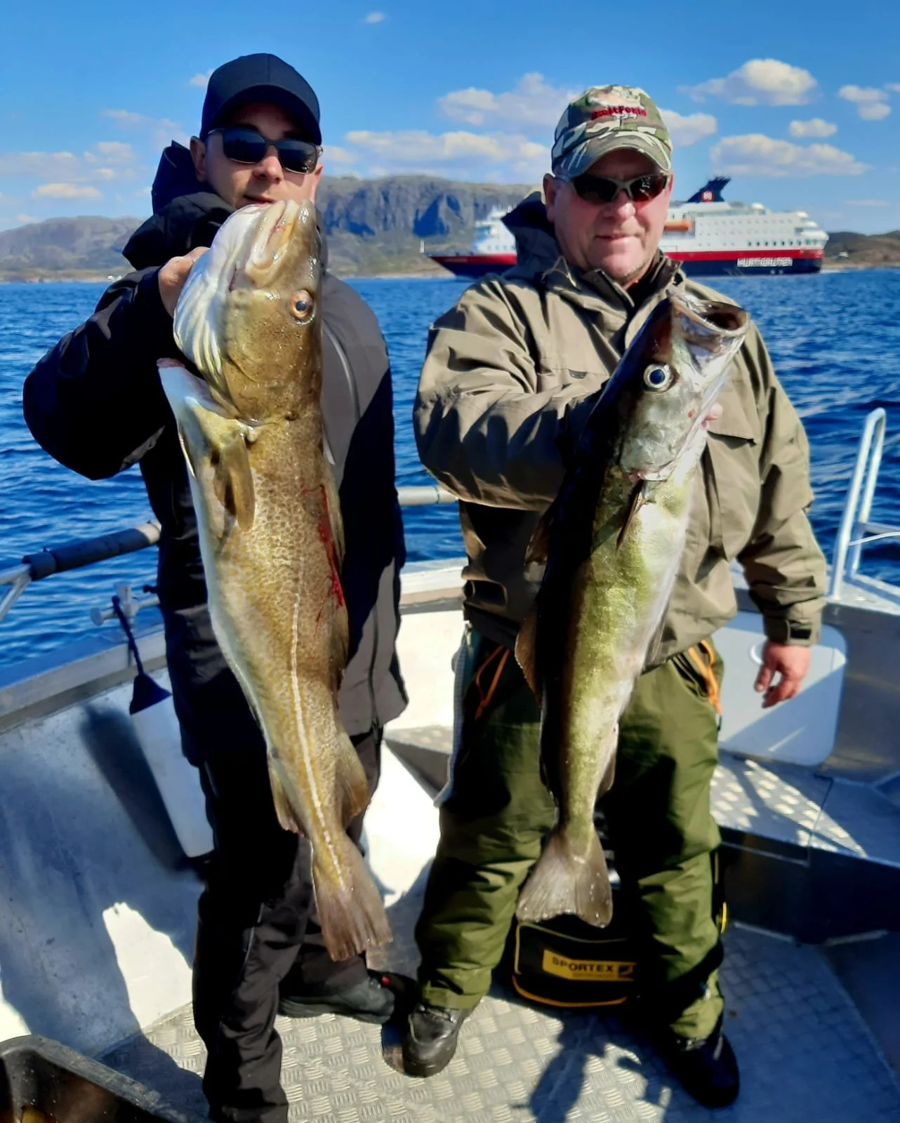 Zwei Männer auf einem Boot, die große Fische halten, vor einer Küstenlandschaft mit Bergen und einem Kreuzfahrtschiff im Wasser.