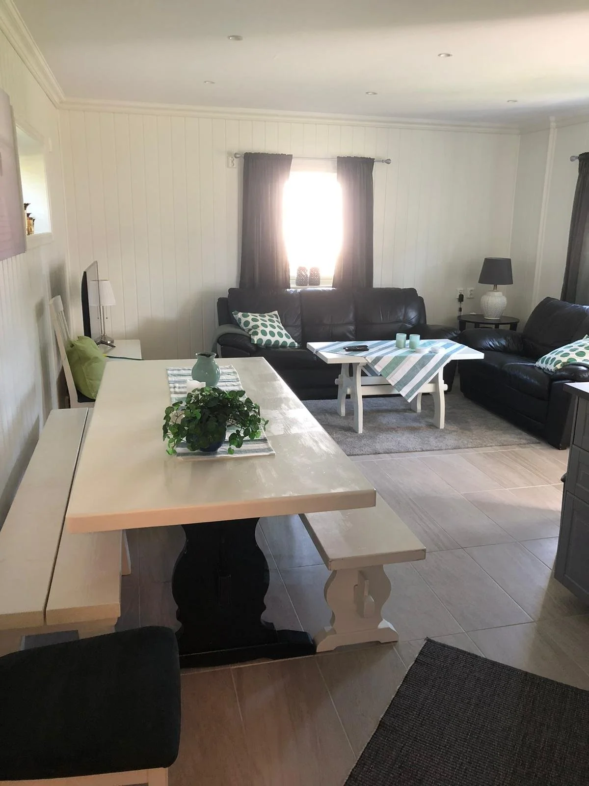 Ein Wohnzimmer mit zwei schwarzen Ledersofas, weißen Kaffeetischen, Vorhängen vor einem Fenster, und Dekorationsgegenständen, darunter Kerzen und Kissen.