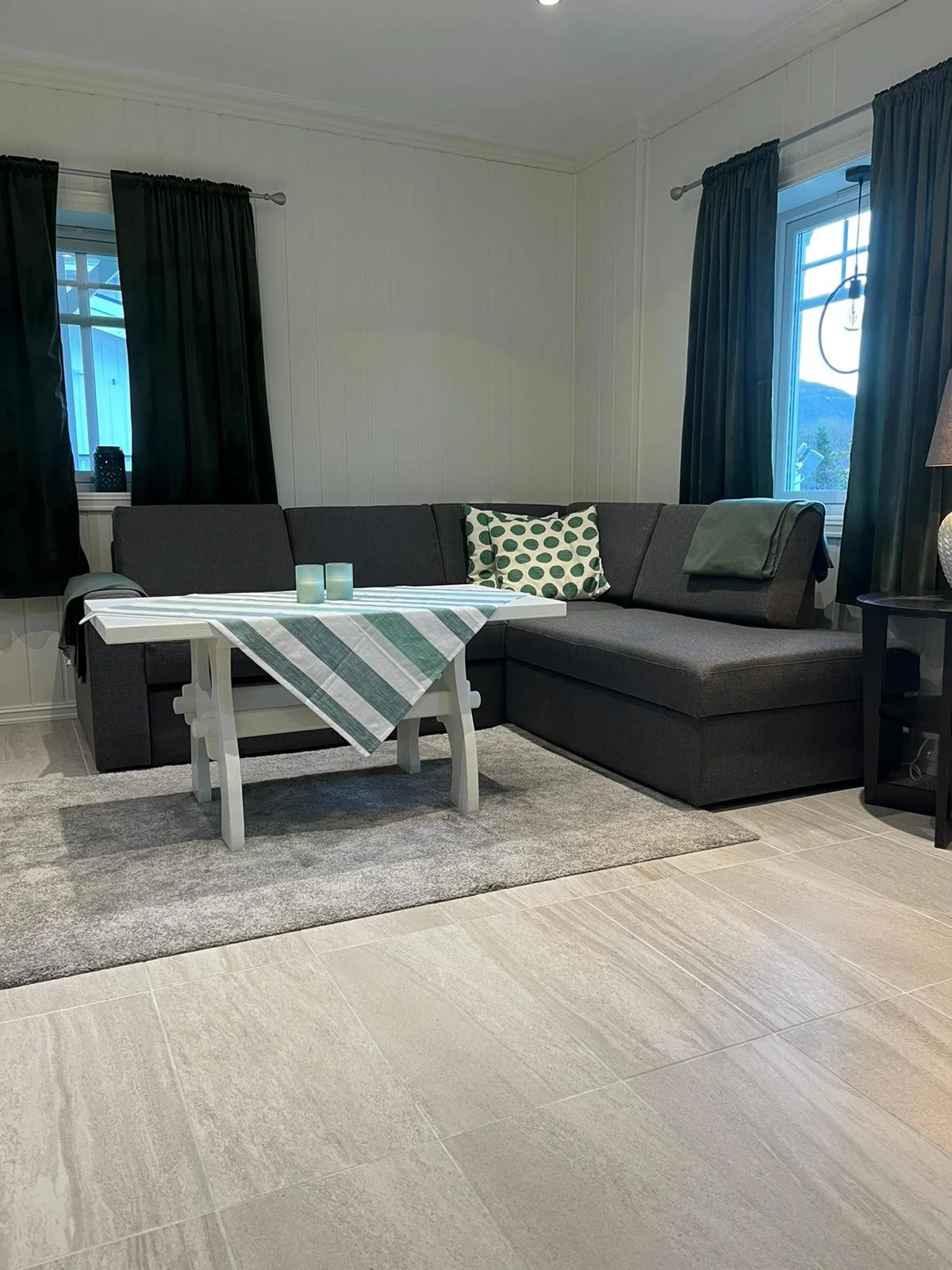 Wohnzimmer mit grauem Sofa, zwei Fenster mit blauen Vorhängen, Kissen und einer Schreibtisch mit Tischdecke und drei Kerzen