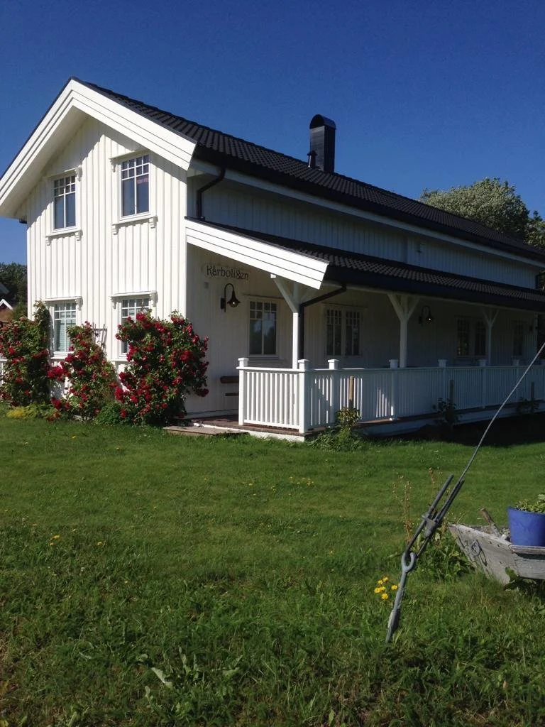 Ein weißes Haus mit dunklem Dach, Vordach und Garten mit Blumen und Rasen, beschriftet mit 'Kärbolsen'.