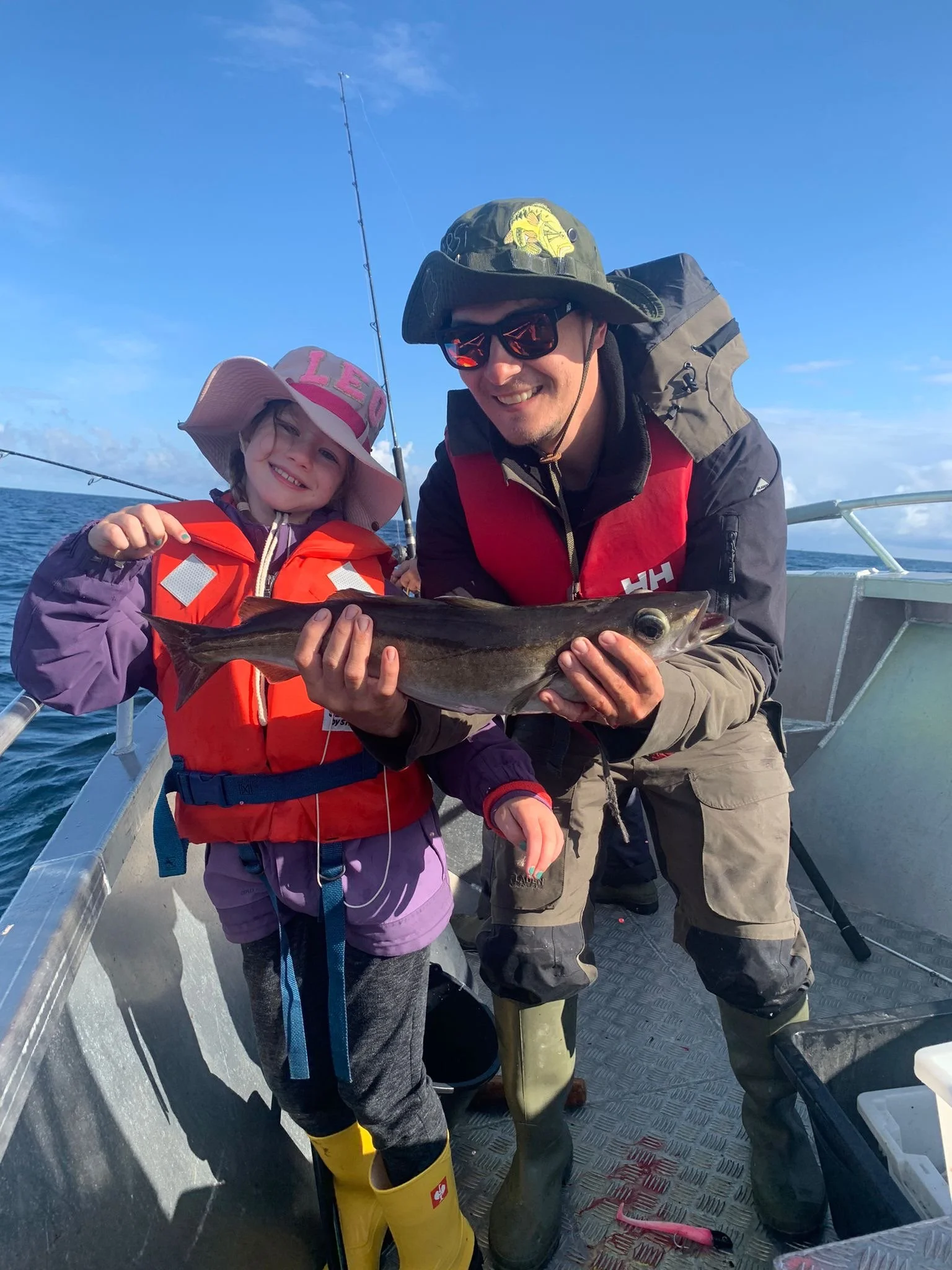 Ein Mann und ein Kind auf einem Boot halten ein Fisch in den Händen, beide tragen Regenkleidung und Schwimmstiefel, das Wetter ist sonnig und der Himmel ist blau.