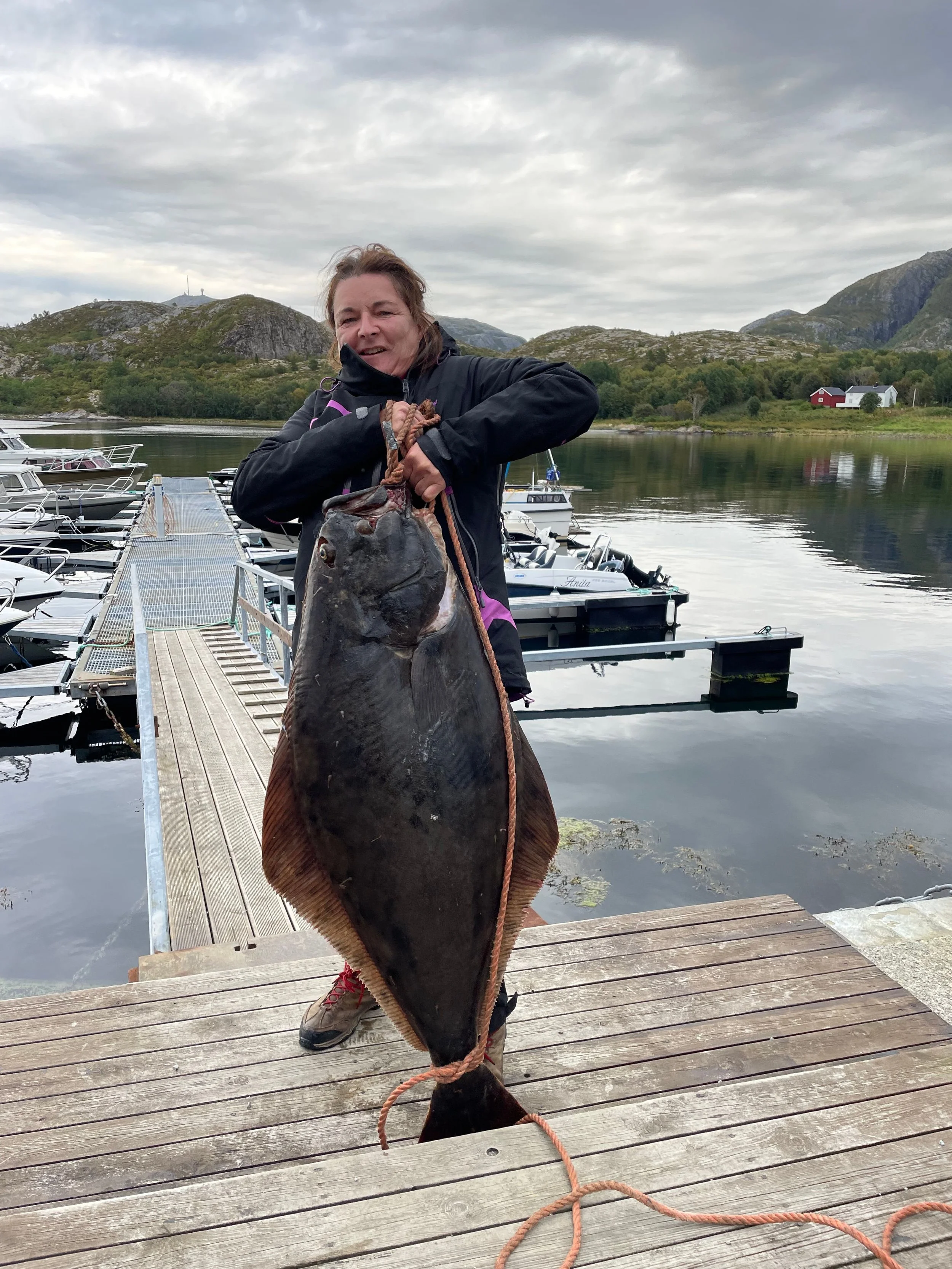 Frau hält über den Steg einen großen Fisch vor einer Marina mit Booten, gegen eine malerische Kulisse von Bergen und See in Norwegen.