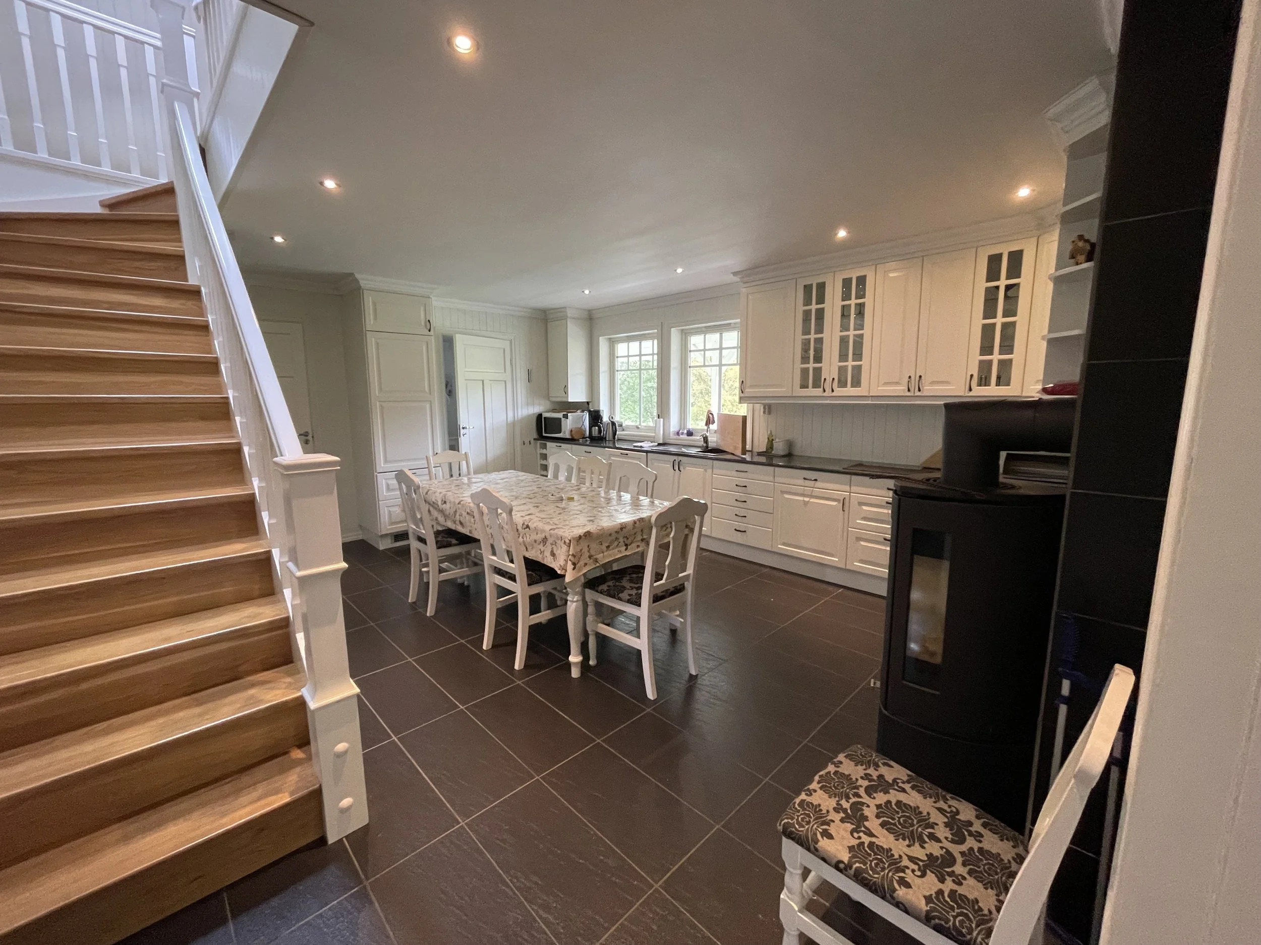 Ein Blick in eine helle, modern eingerichtete Küche mit weißen Küchenmöbeln, einem Esstisch mit Blumenmuster-Tischdecke und schwarzen Fliesen auf dem Boden. Im Hintergrund große Fenster, die natürliches Licht hereinlassen, und eine Treppe links im Bi