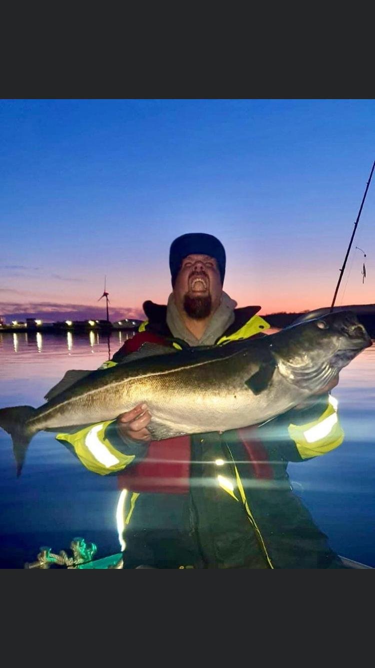 Ein Mann hält einen großen Fisch beim Angeln im Wasser bei Sonnenuntergang, im Hintergrund sind Windräder und der Himmel ist blau und orange.