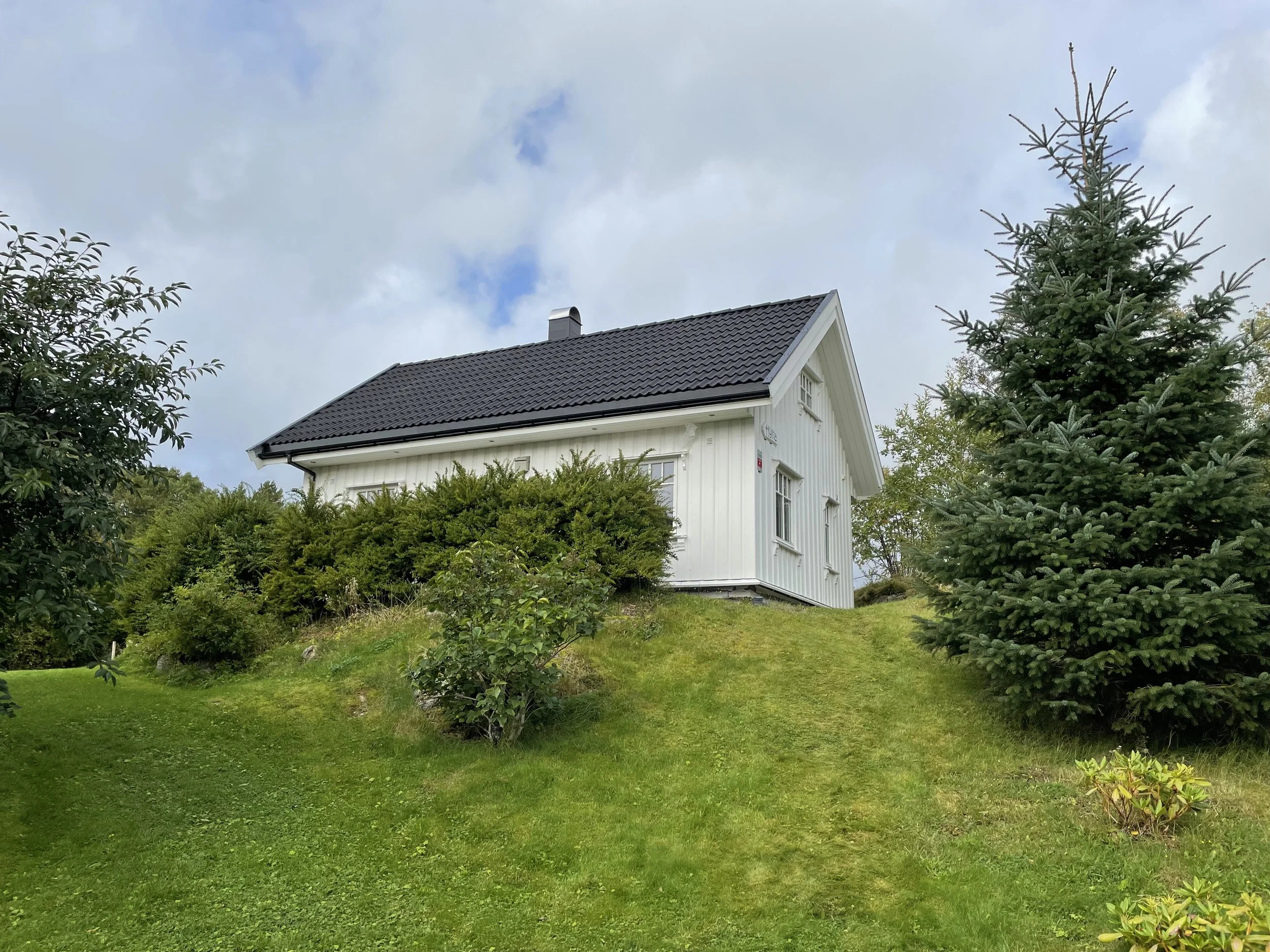 Ein weißes Haus mit grauem Dach, umgeben von grünen Büschen und Nadelbäumen, unter einem bewölkten Himmel.