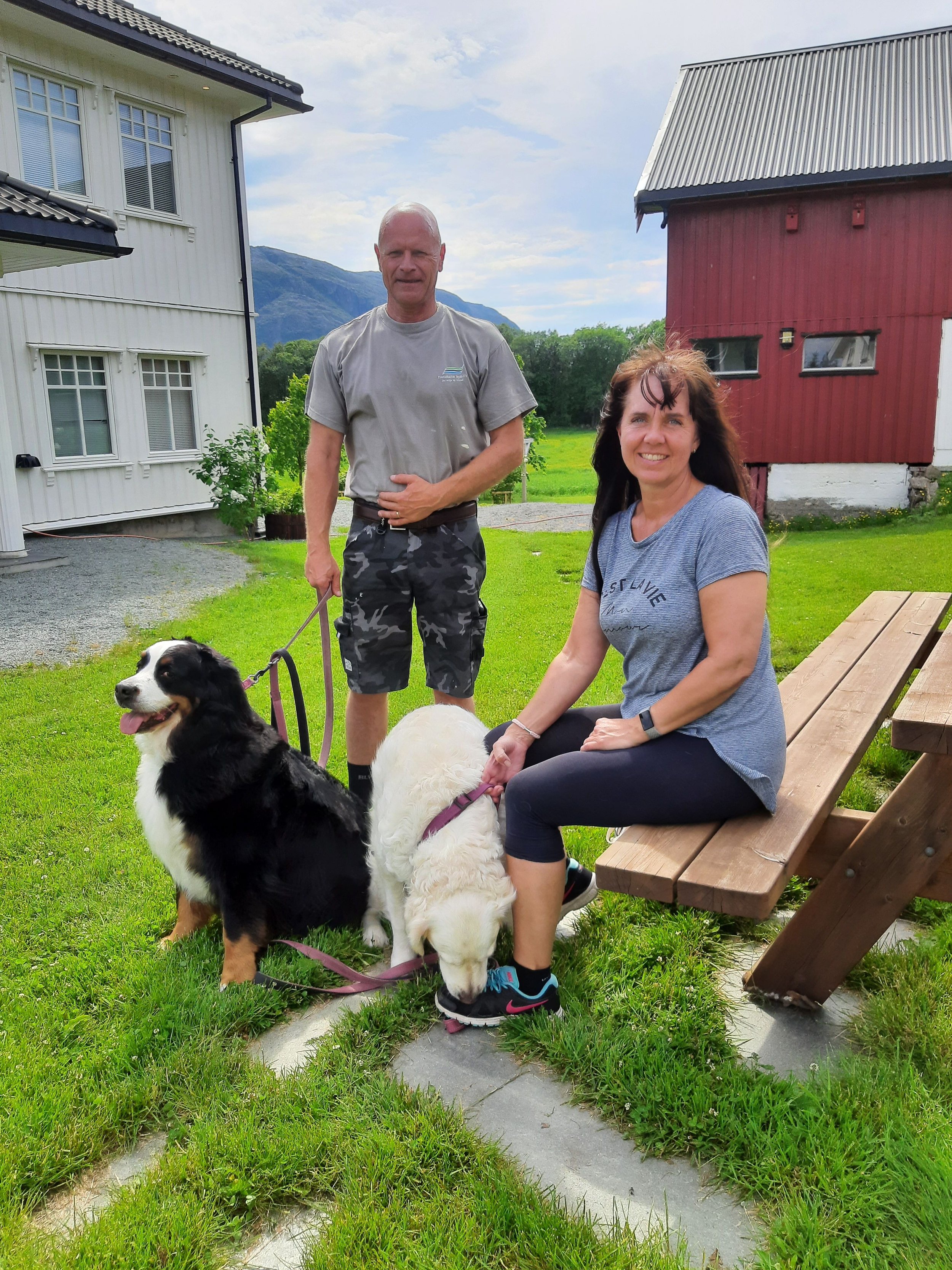 Ein Mann und eine Frau sitzen und stehen mit zwei Hunden auf einer Wiese vor Häusern in einer ländlichen Umgebung.
