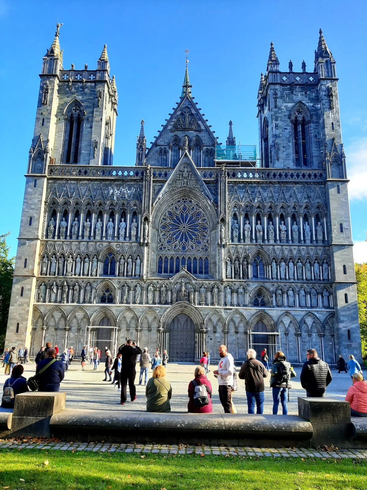 Die Fassade der Kathedrale Notre-Dame in Paris mit Menschen vor dem Eingang bei Sonnenlicht, blauer Himmel, gotische Architektur mit Türmen und Fenstern.