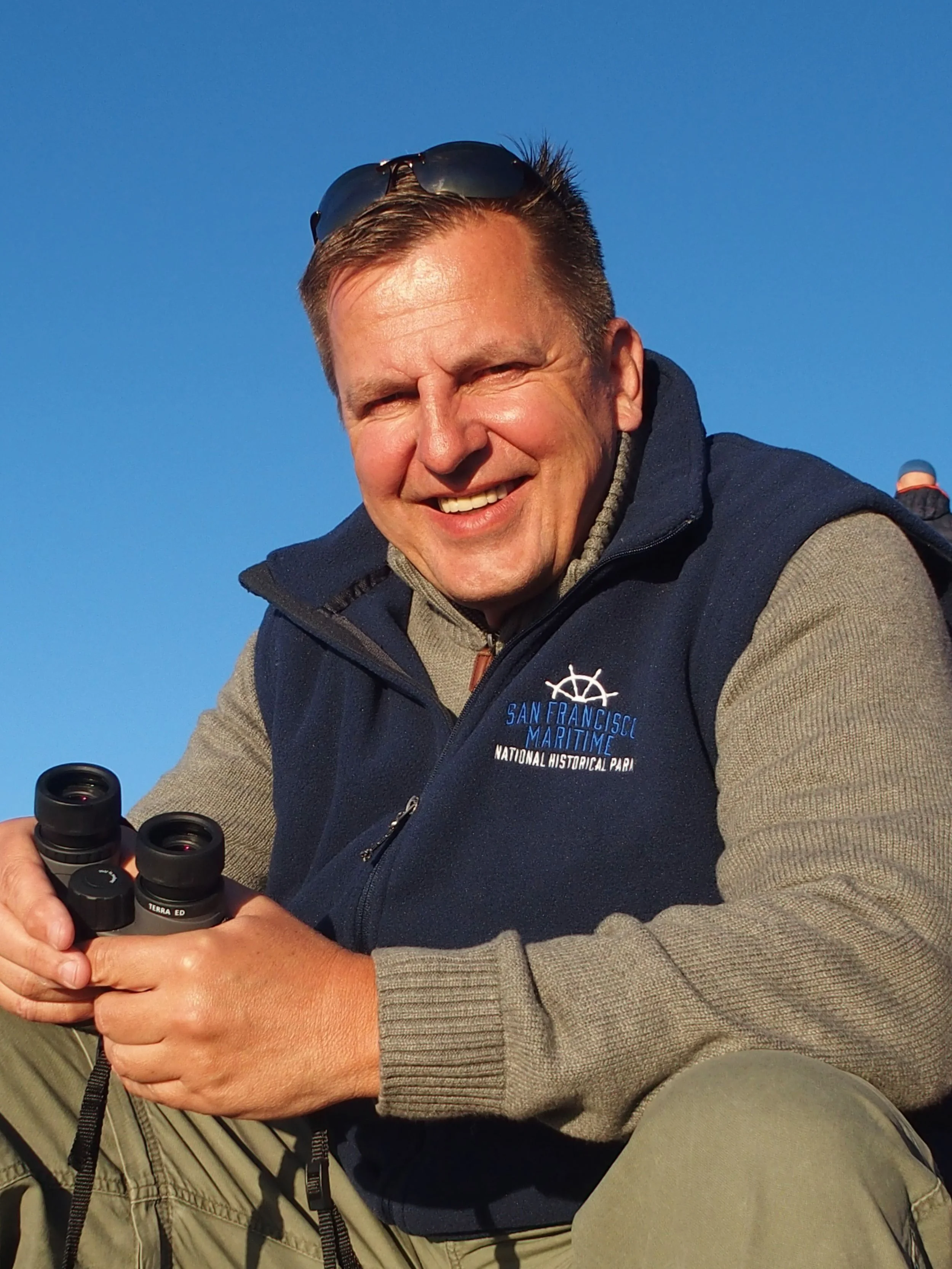 Lächelnder Mann mit Sonnenbrille auf dem Kopf, der mit einem Fernglas in den Händen auf einem Berg sitzt, bei Sonnenlicht unter blauem Himmel.