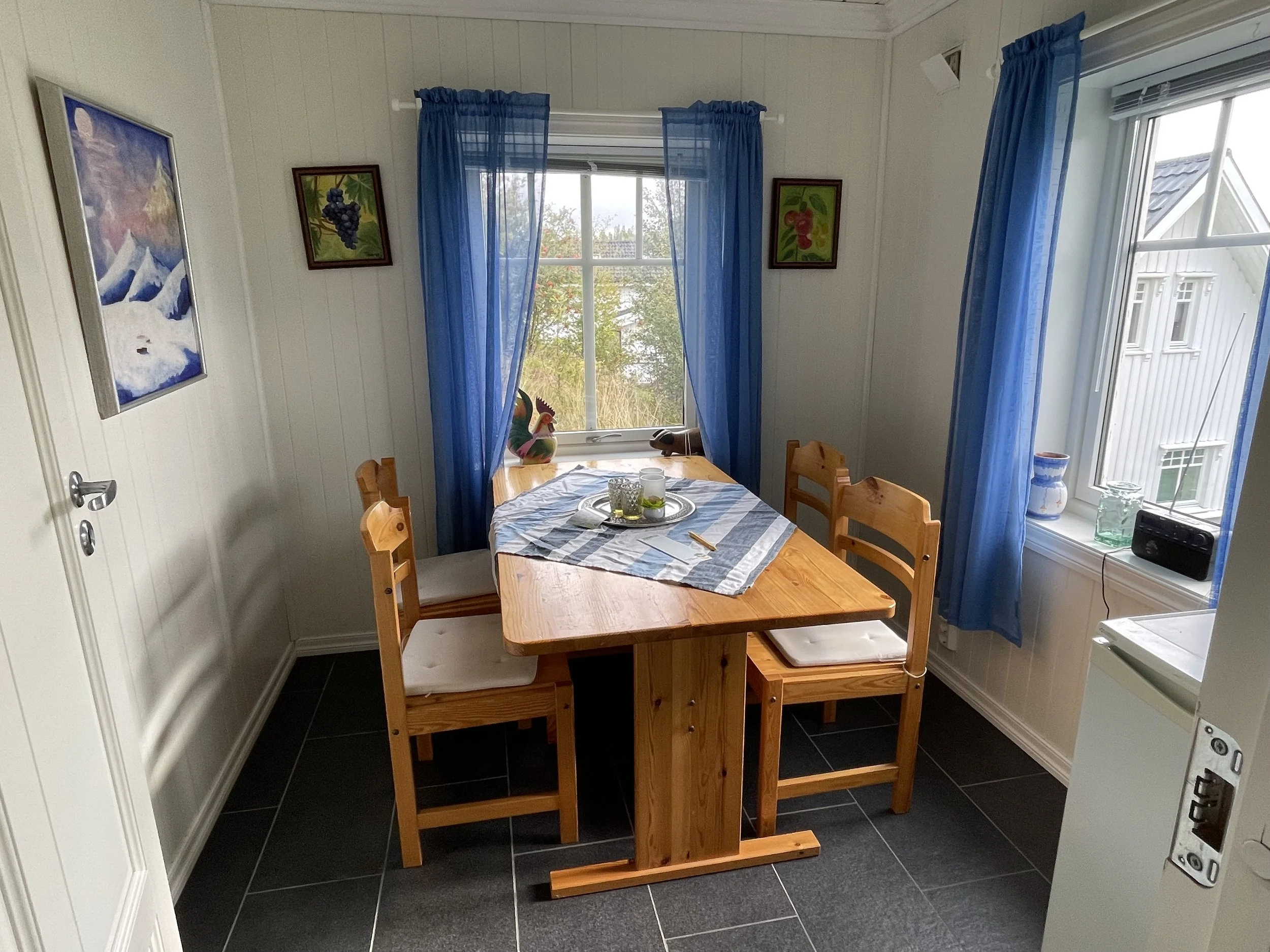 Ein Esszimmertisch mit sechs Stühlen in einem Raum mit weißen Wänden, blauen Vorhängen vor den Fenstern und Wandbildern mit Bergen und Trauben.