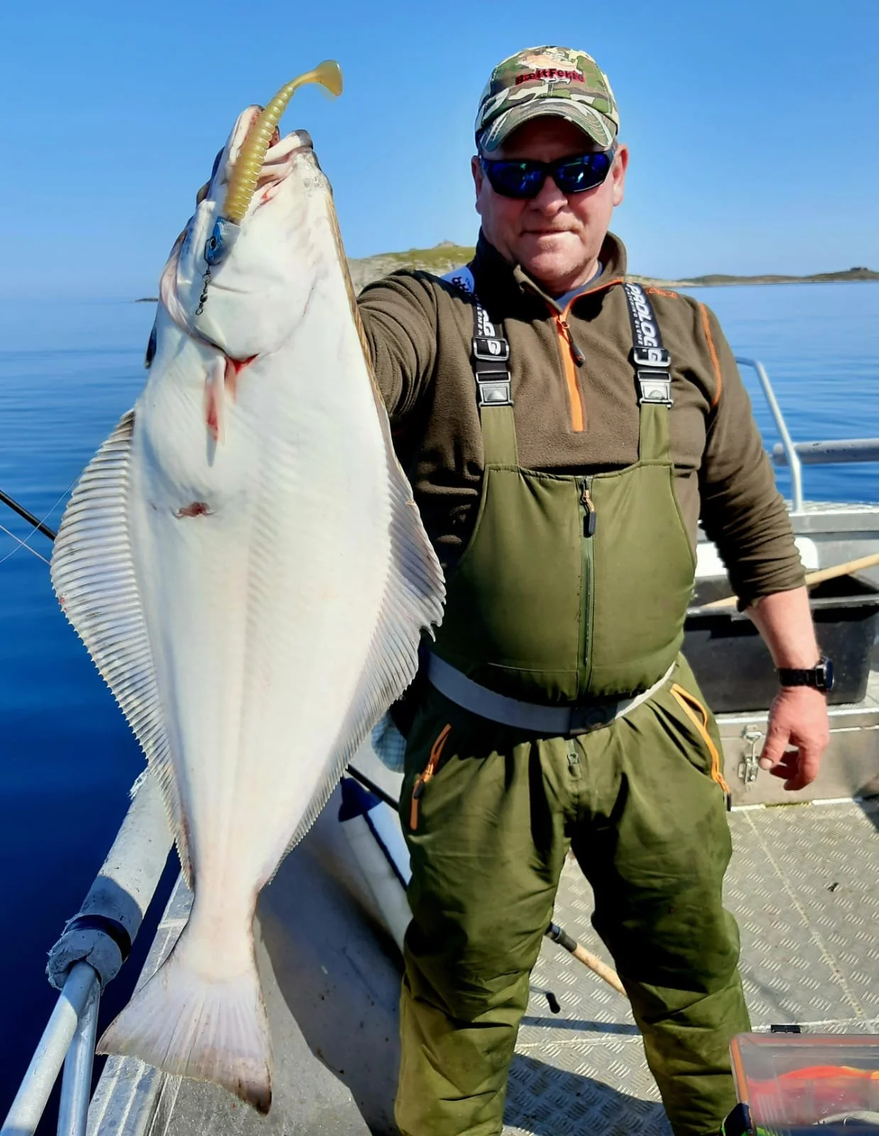 Mann in Angelbekleidung hält einen großen Fisch mit einer Angel auf einem Boot auf dem Wasser bei schönem Wetter.
