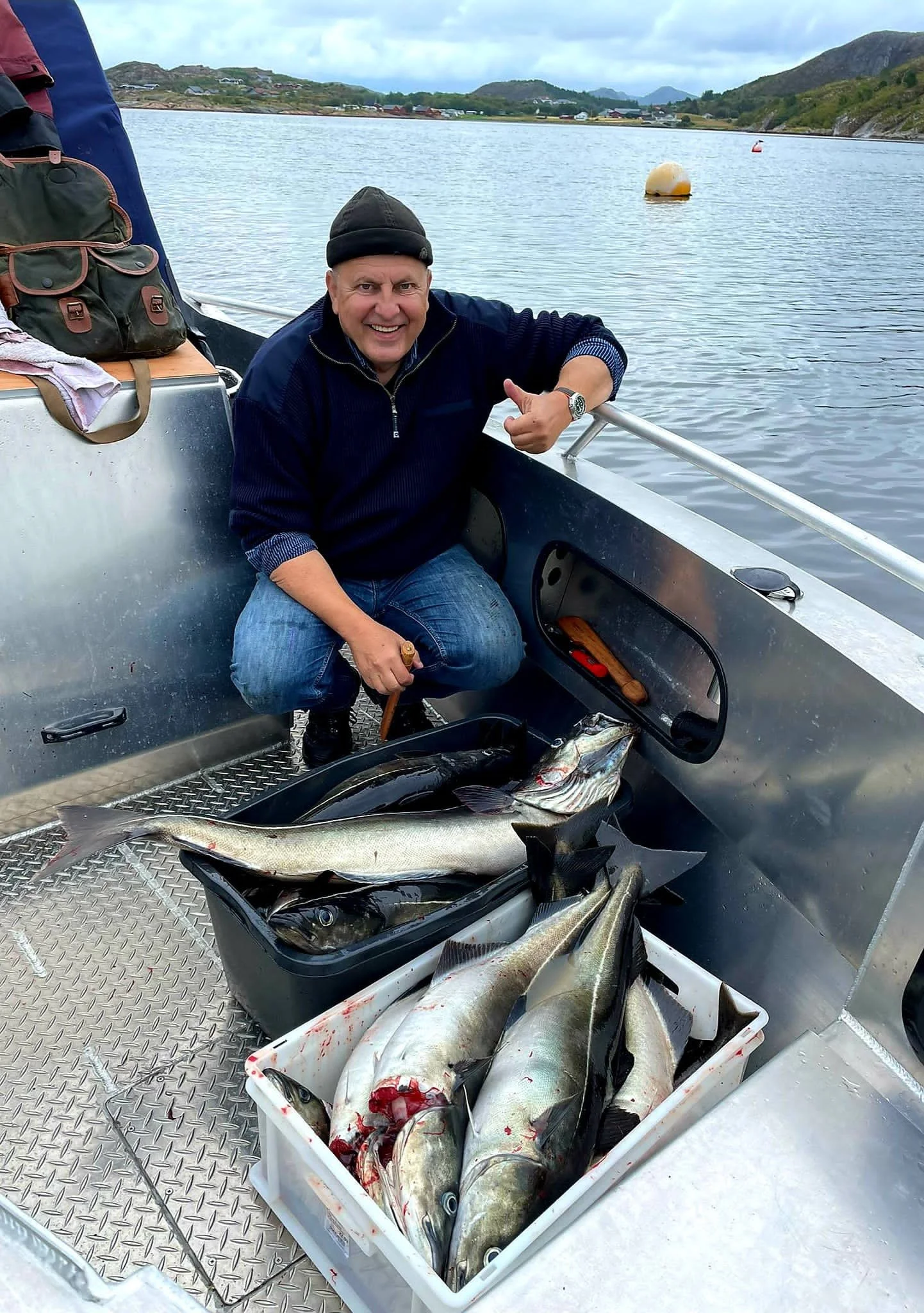 Ein glücklicher älterer Mann auf einem Boot, zeigt auf die gefangenen Fische, die in Behältern vor ihm liegen, mit einer See- oder Flusslandschaft im Hintergrund.