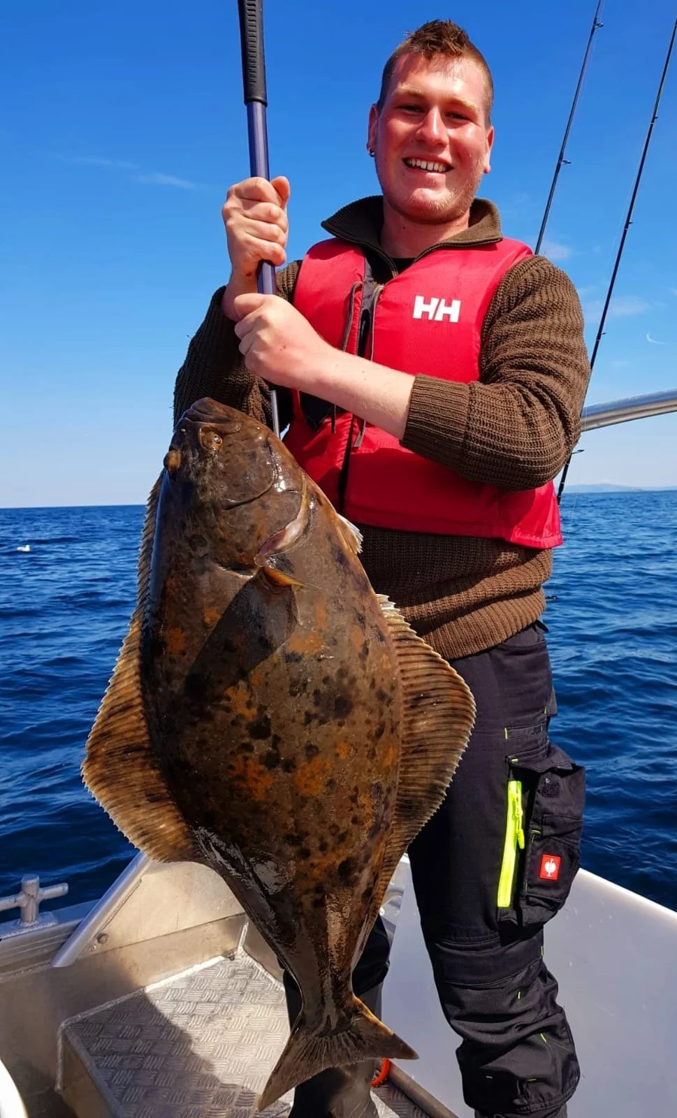 Mann auf einem Boot hält einen großen Fisch, der vermutlich ein Plattfisch ist, mit einem Angelsec, im Hintergrund das Meer und blauer Himmel.