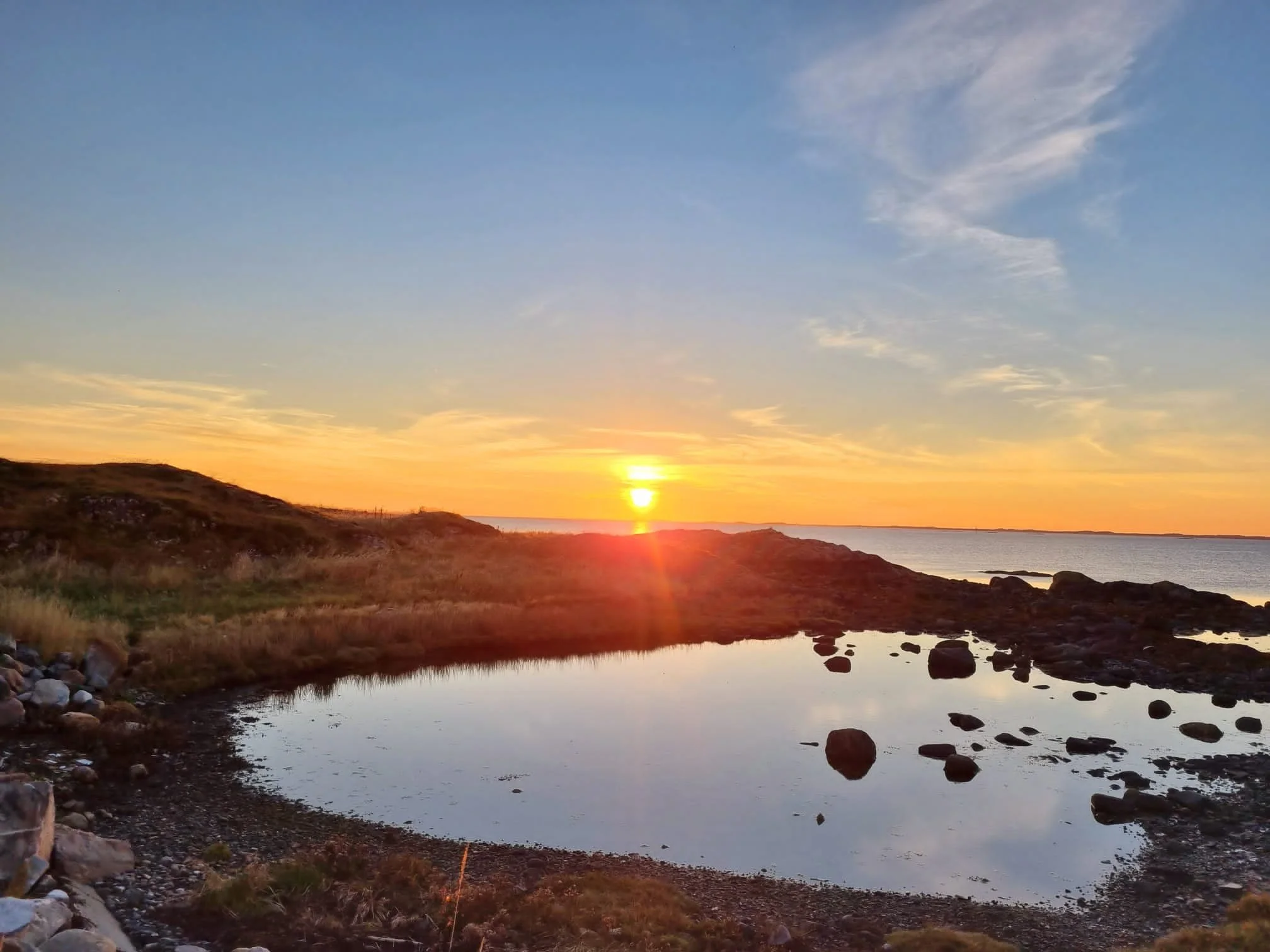Der Sonnenuntergang über einer Küstenlandschaft mit Wasserbecken, Steinen und Ufervegetation.