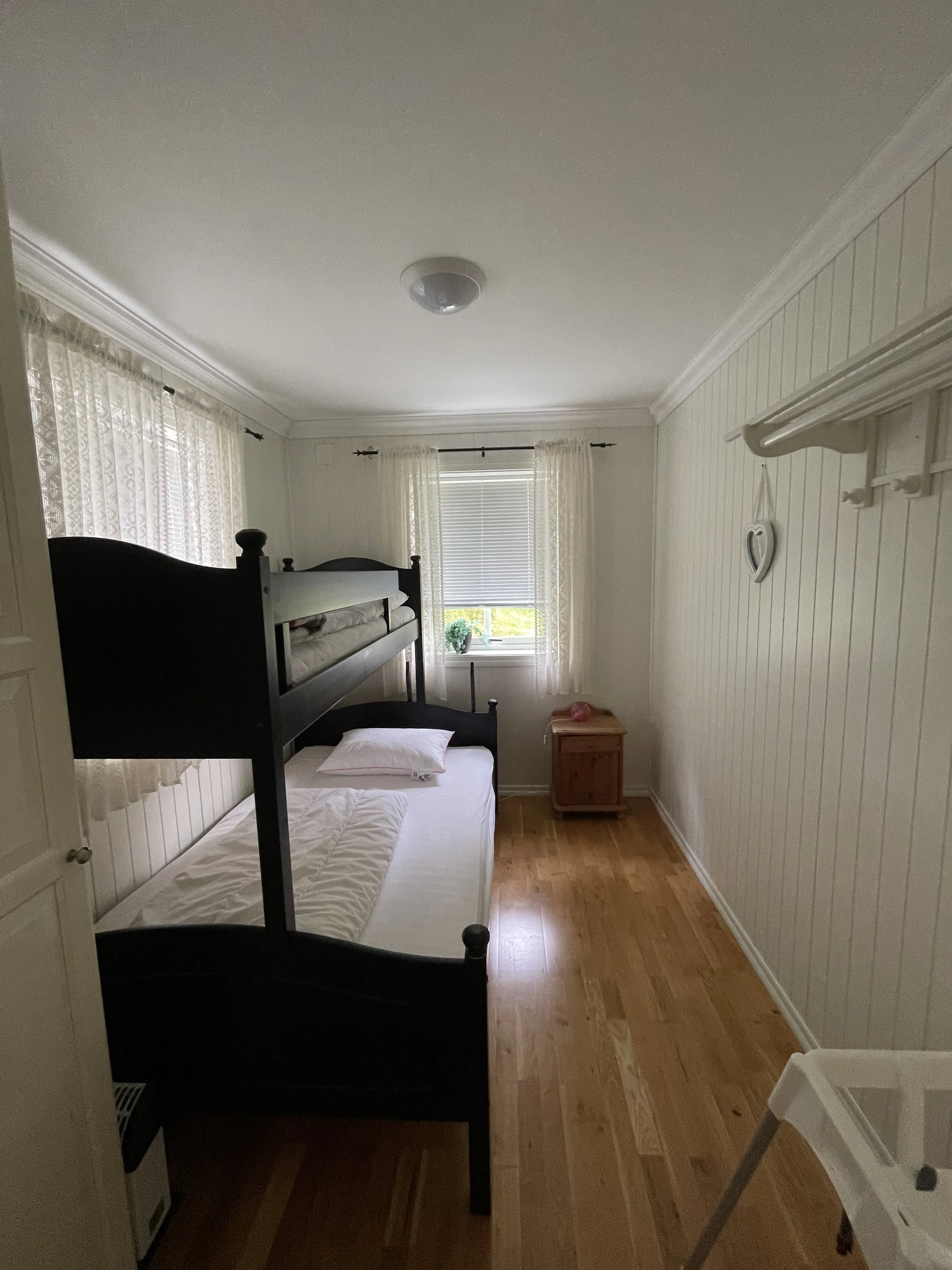 Ein Schlafzimmer mit Holzboden, weißen Wänden und Vorhängen vor Fenstern. Es gibt ein Hochbett und einen kleinen Nachttisch. Ein Fenster mit Sicht nach draußen ist sichtbar, sowie eine weiße Wanddekoration in Herzform. Ein Kindersitz ist im Vordergru