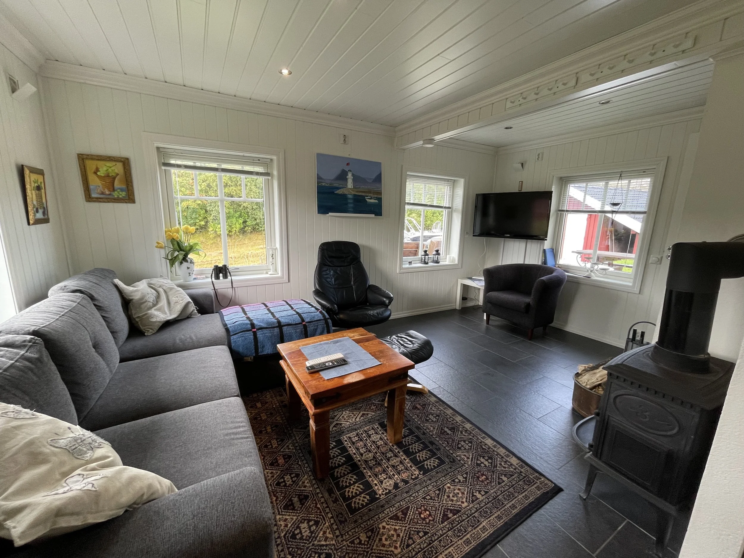 Ein Wohnzimmer mit grauer Couch, zwei schwarzen Sesseln, einem Couchtisch aus Holz, einem Teppich mit Muster, einem Fernseher, mehreren Fenstern und Kunstwerken an den Wänden.