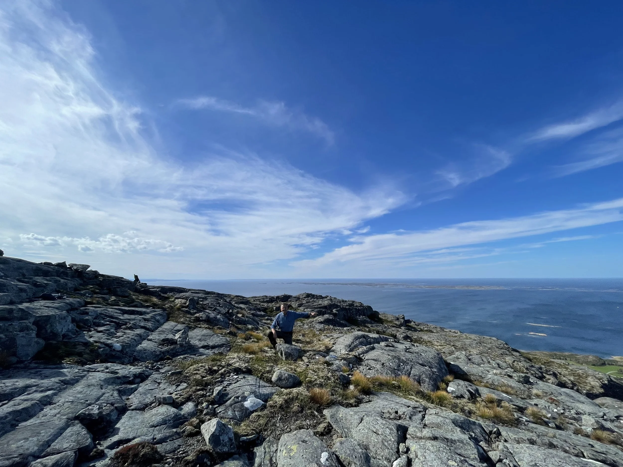 Küstenlandschaft mit Felsen, ein Mann kniet auf einem Felsen, blauer Himmel mit Wolken, Blick auf das Meer und kleine Inseln im Hintergrund.