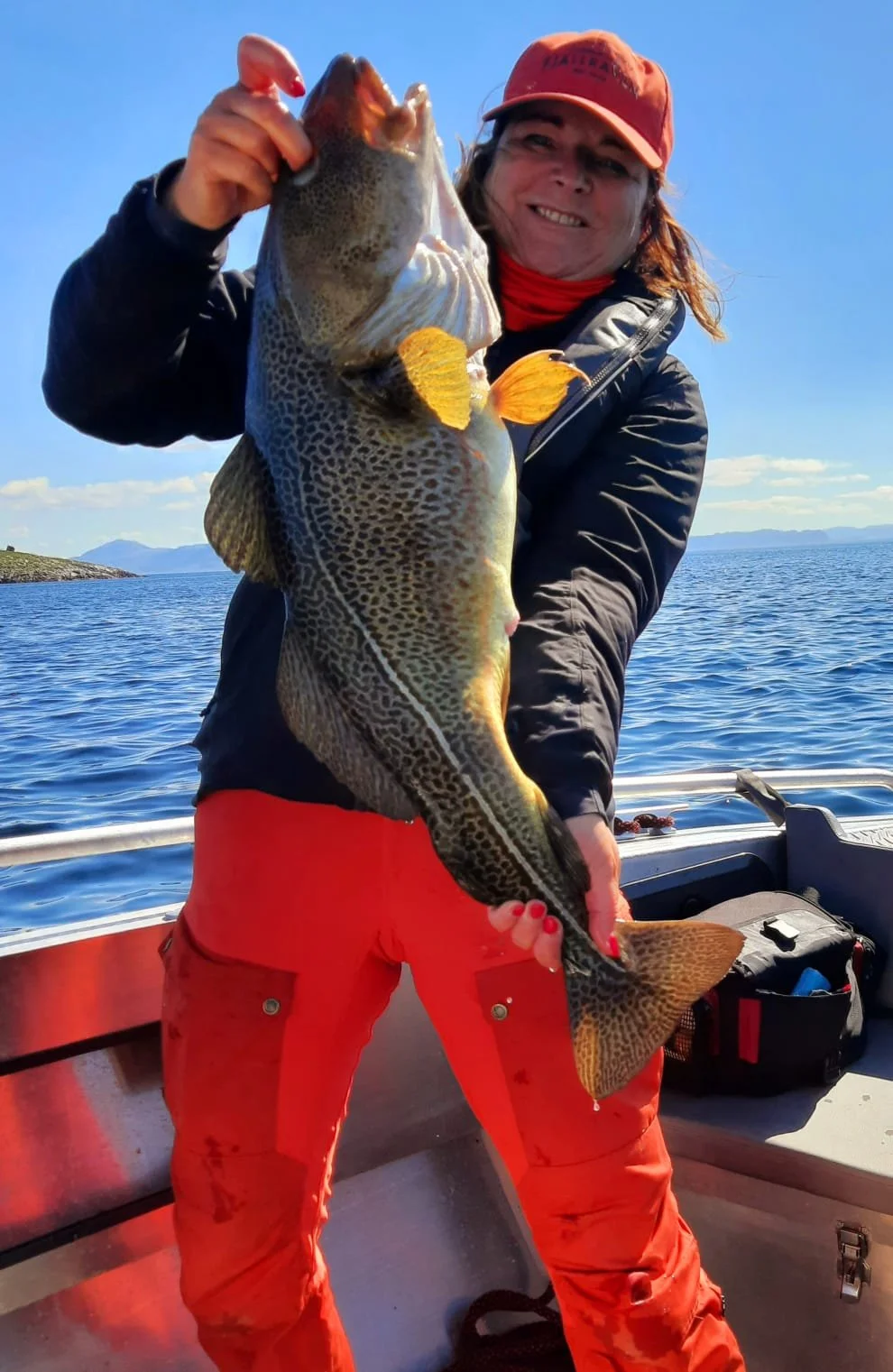 Frau mit roter Mütze und roten Hosen hält einen großen Fisch auf einem Boot vor einem blauen Himmel und Wasser