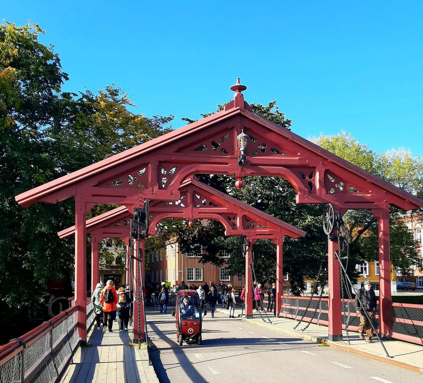 Eine Brücke mit roten Holzkonstruktionen und Schienen, auf der viele Menschen gehen, darunter auch ein kleines rotes Elektrofahrzeug.